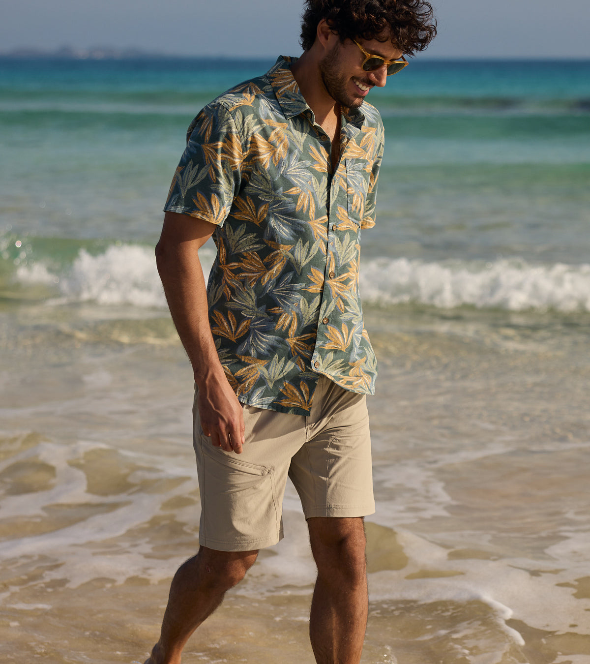 A smiling man with brown hair walks along the beach in the sun. The man is wearing a green shirt with yellow and blue palm leaves printed on top.