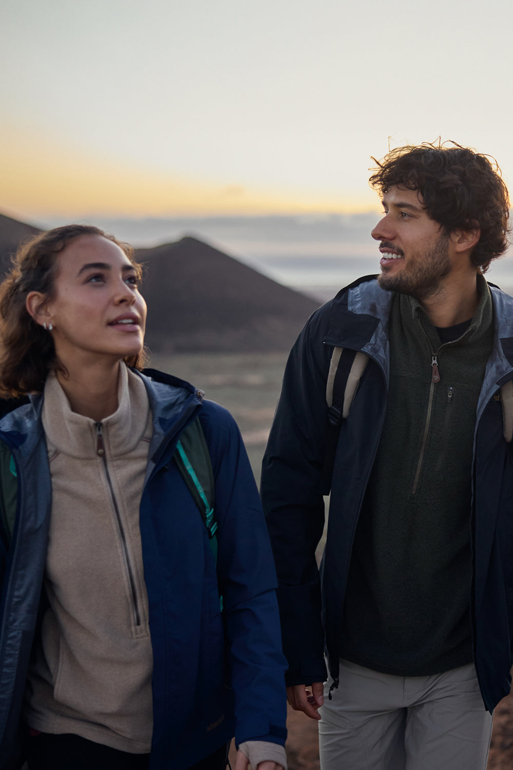A woman and man, wearing blue rain coats and fleeces, walk on a mountain path at sunrise.  The sun is rising over the mountain behind them.