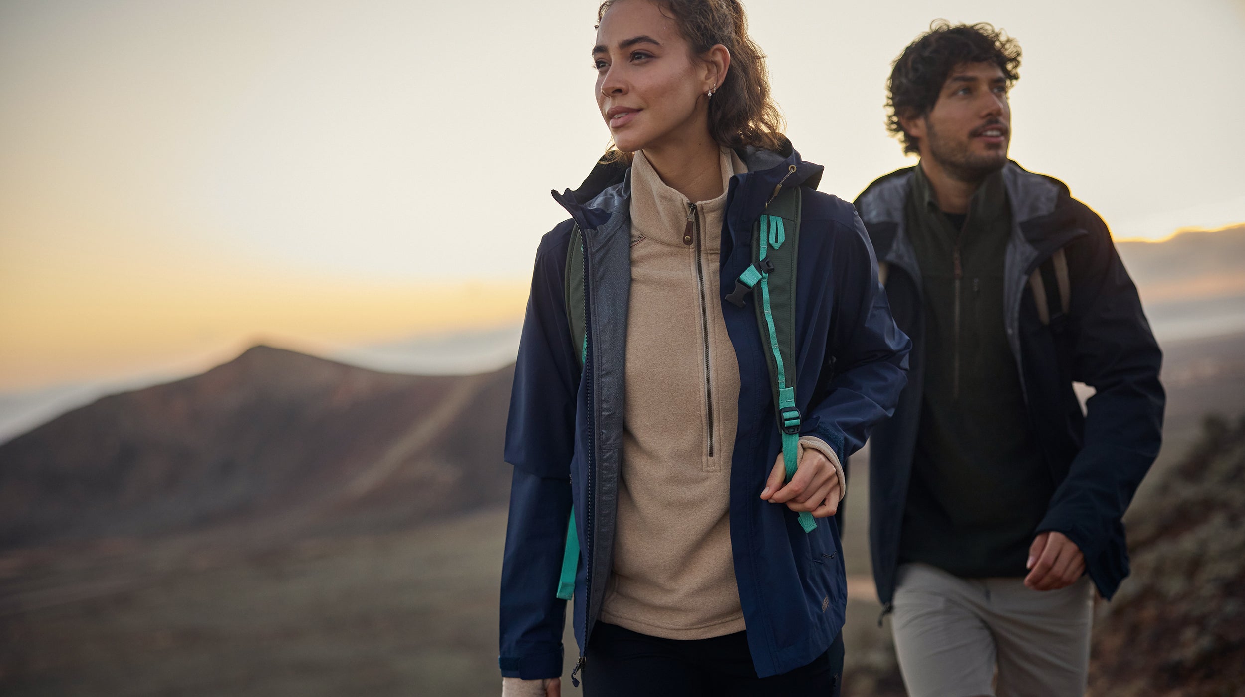 A woman and man, wearing blue rain coats and fleeces, walk on a mountain path at sunrise.  The sun is rising over the mountain behind them.