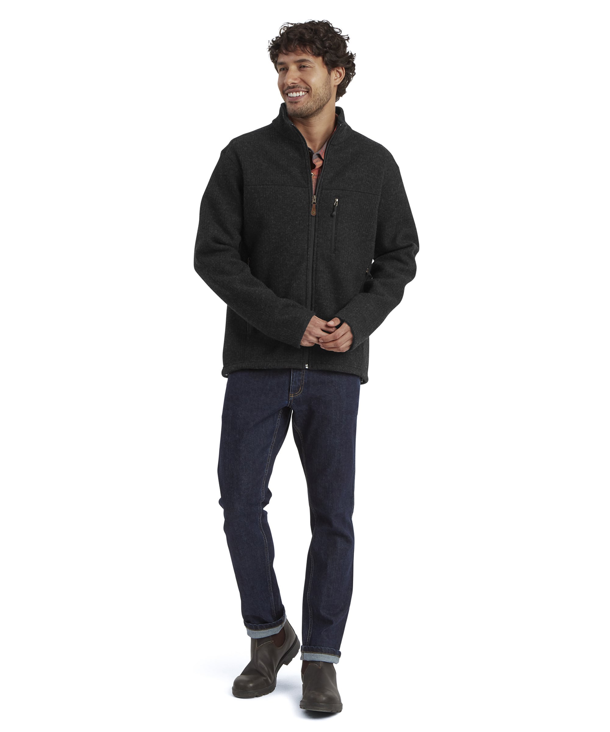 A full length image of a  man wearing a black wool fleece jacket standing against a white background. He is smiling at the camera with both hands together. The jacket has a full length zip with a leather puller and a small, zipped chest pocket. The jacket is paired some blue jeans and brown leather boots.