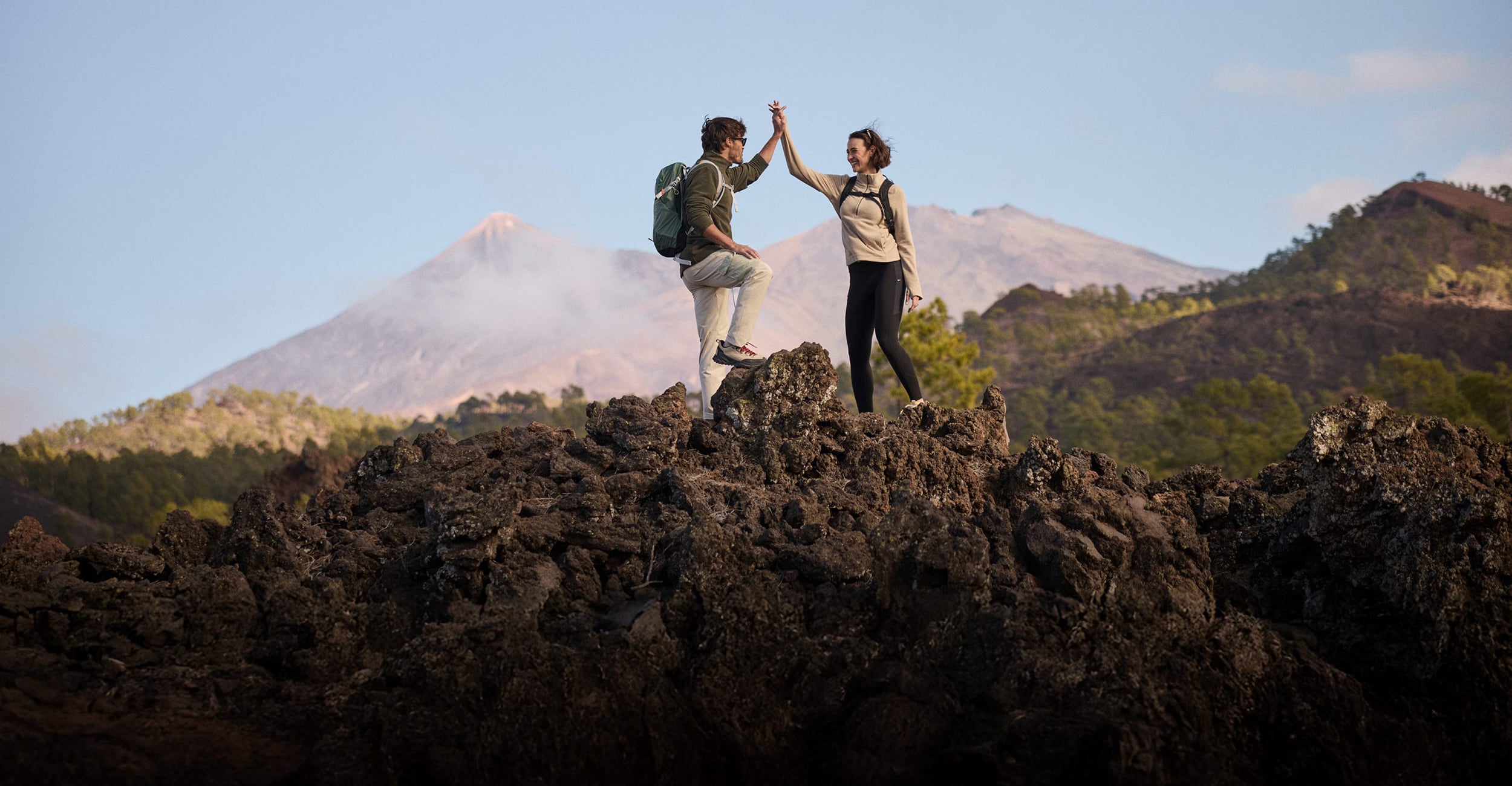 Two hikers standing on dark volcanic rock in a mountain landscape, high-fiving with a volcano and blue sky in the background.