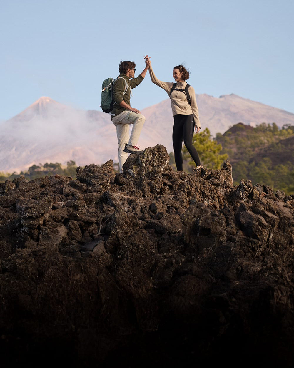 Two hikers standing on dark volcanic rock, high-fiving with mountains in the background under a clear blue sky.