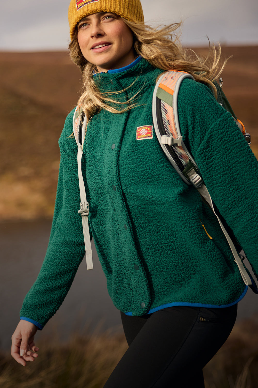 A woman hiking outdoors wearing a green fleece jacket, a yellow beanie, and a backpack, with a brown hillside in the background.