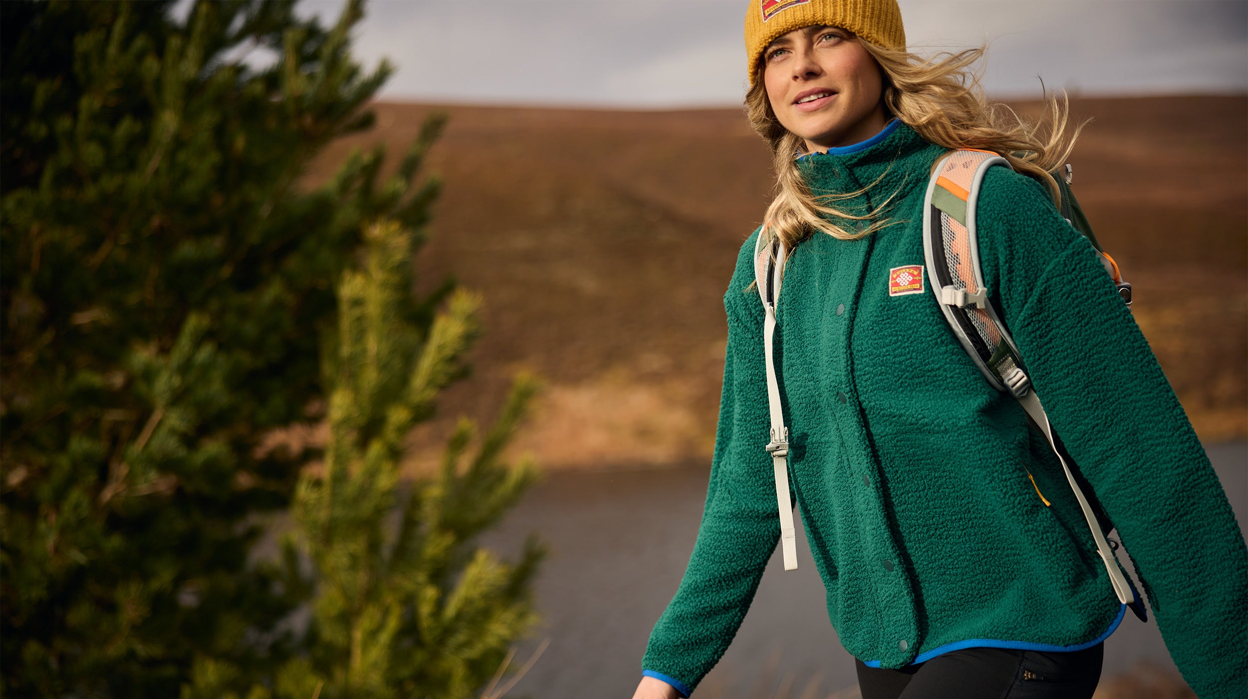 A woman hiking outdoors wearing a green fleece jacket, a yellow beanie, and a backpack, with a brown hillside in the background.