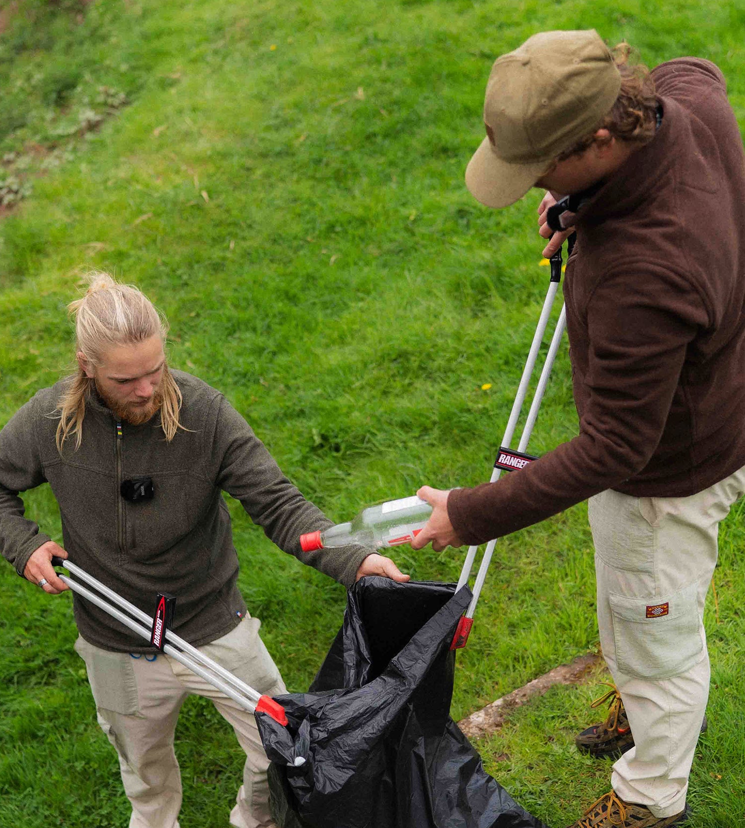 Two young men in a field picking up litter using litter picking tools and a black bin bag. One is wearing a brown fleece and the other a green.