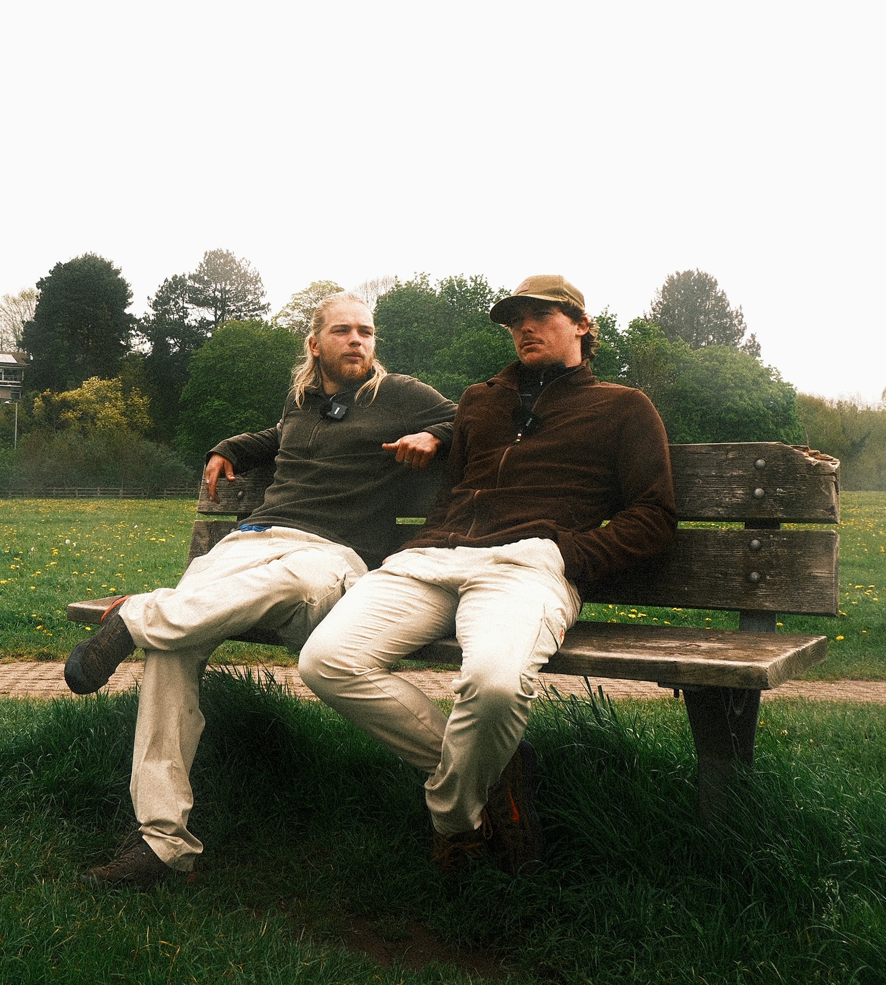 Two men sitting on a wooden bench in a park with trees and grass in the background.