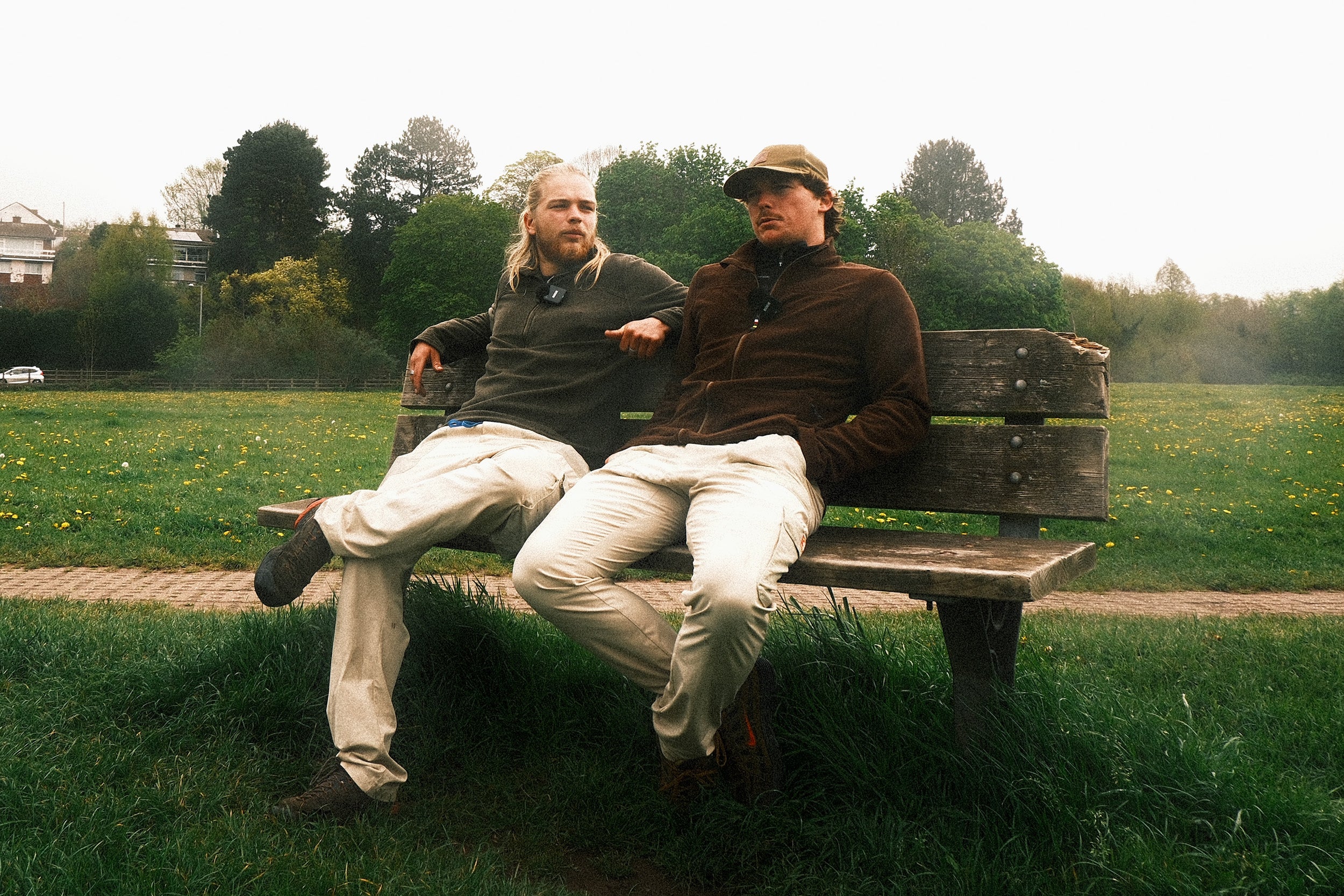 Two men sitting on a wooden bench in a park with trees and grass in the background.
