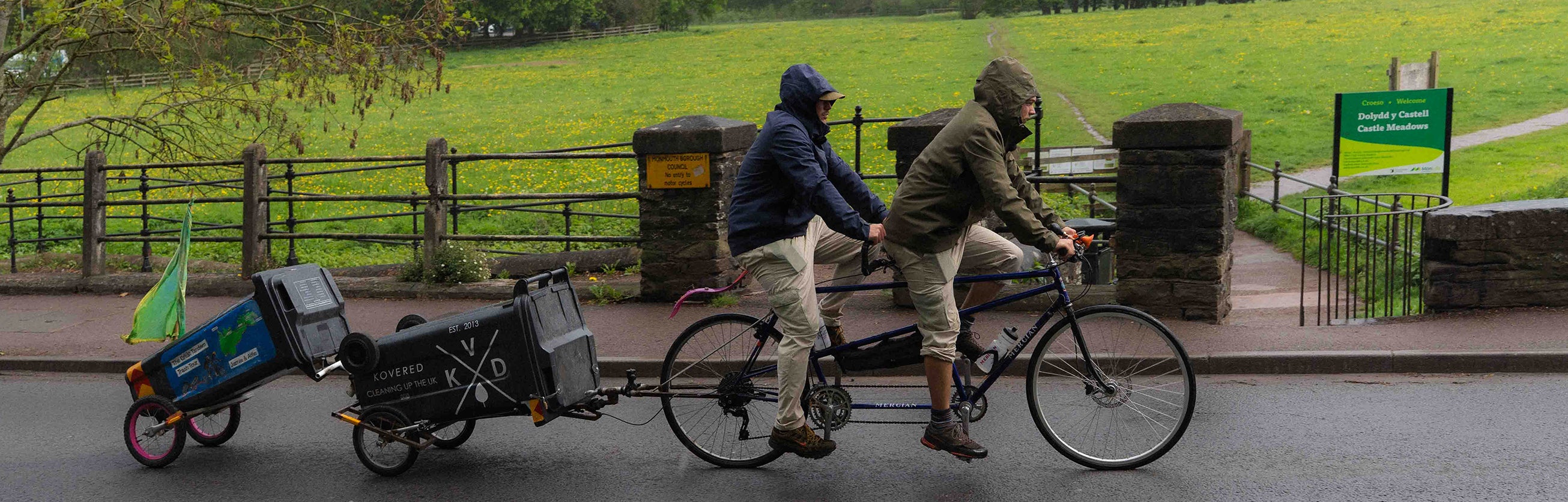 Two men riding a tandem bicycle attached to wheelie bin. They are cycling through a welsh valley, in the rain.