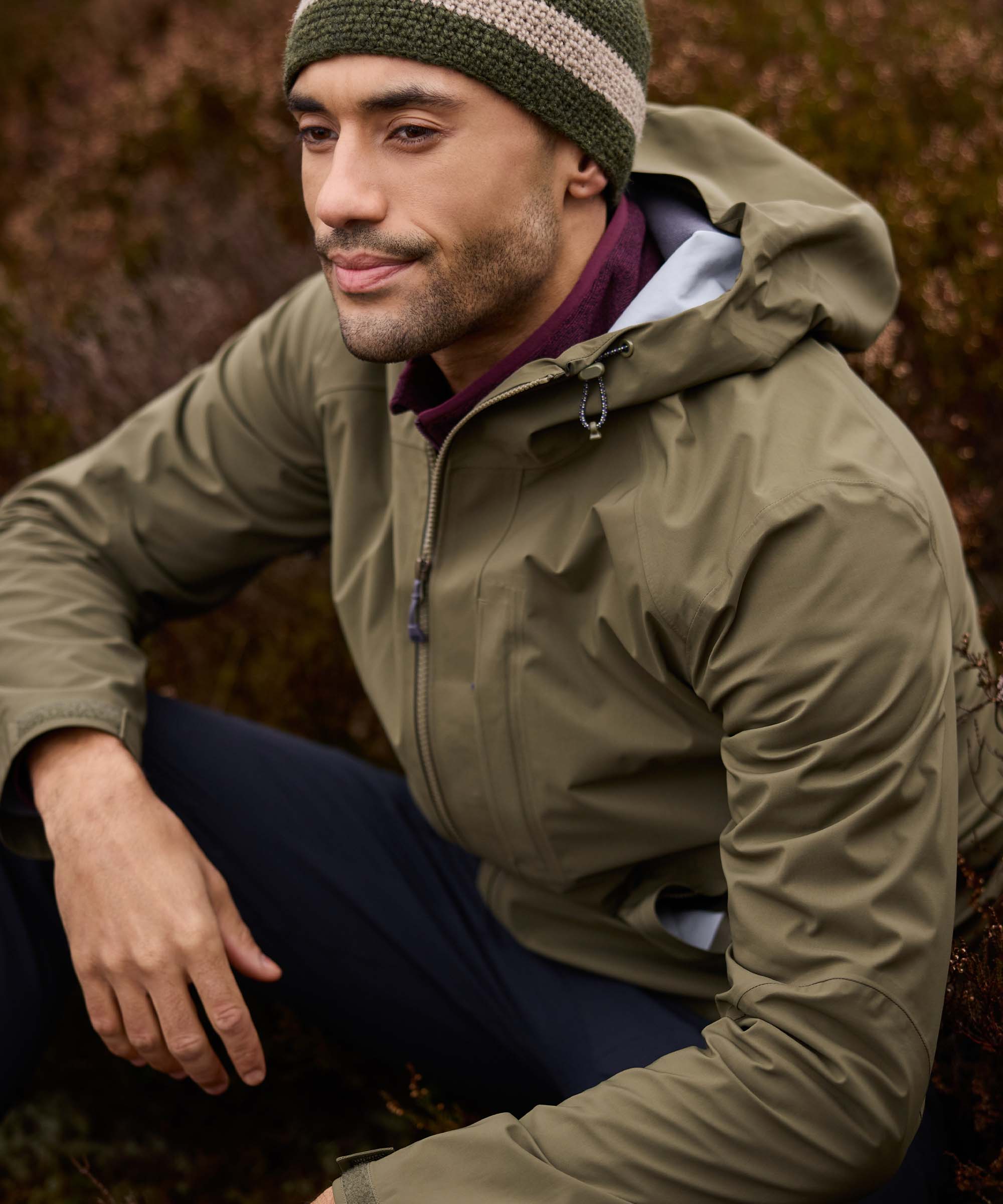 A mid shot of a man wearing a green waterproof jacket and green knit hat sitting outdoors.