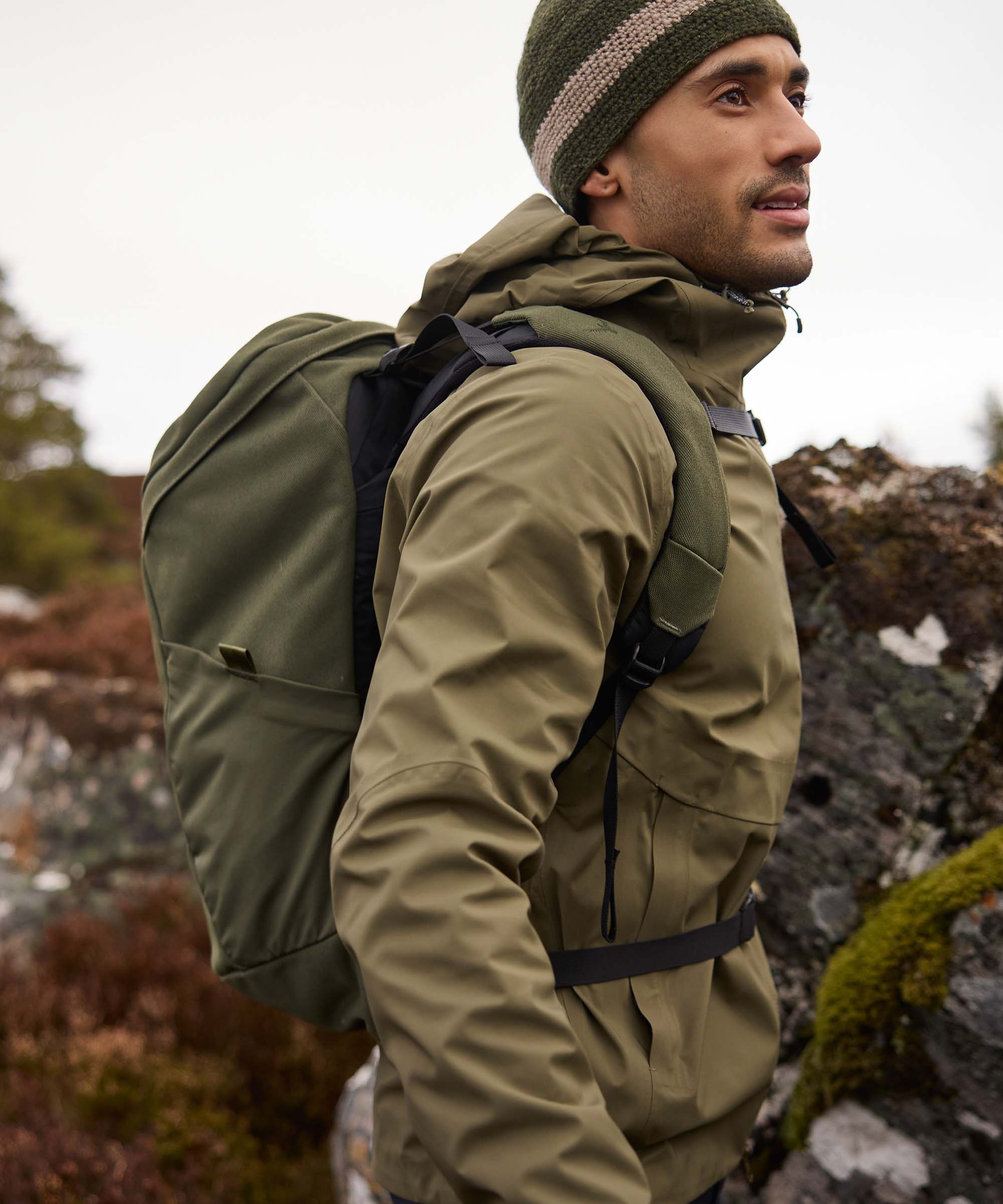A mid shot of a man hiking in a rocky setting whilst wearing a green waterproof jacket, a backpack and green knitted hat.