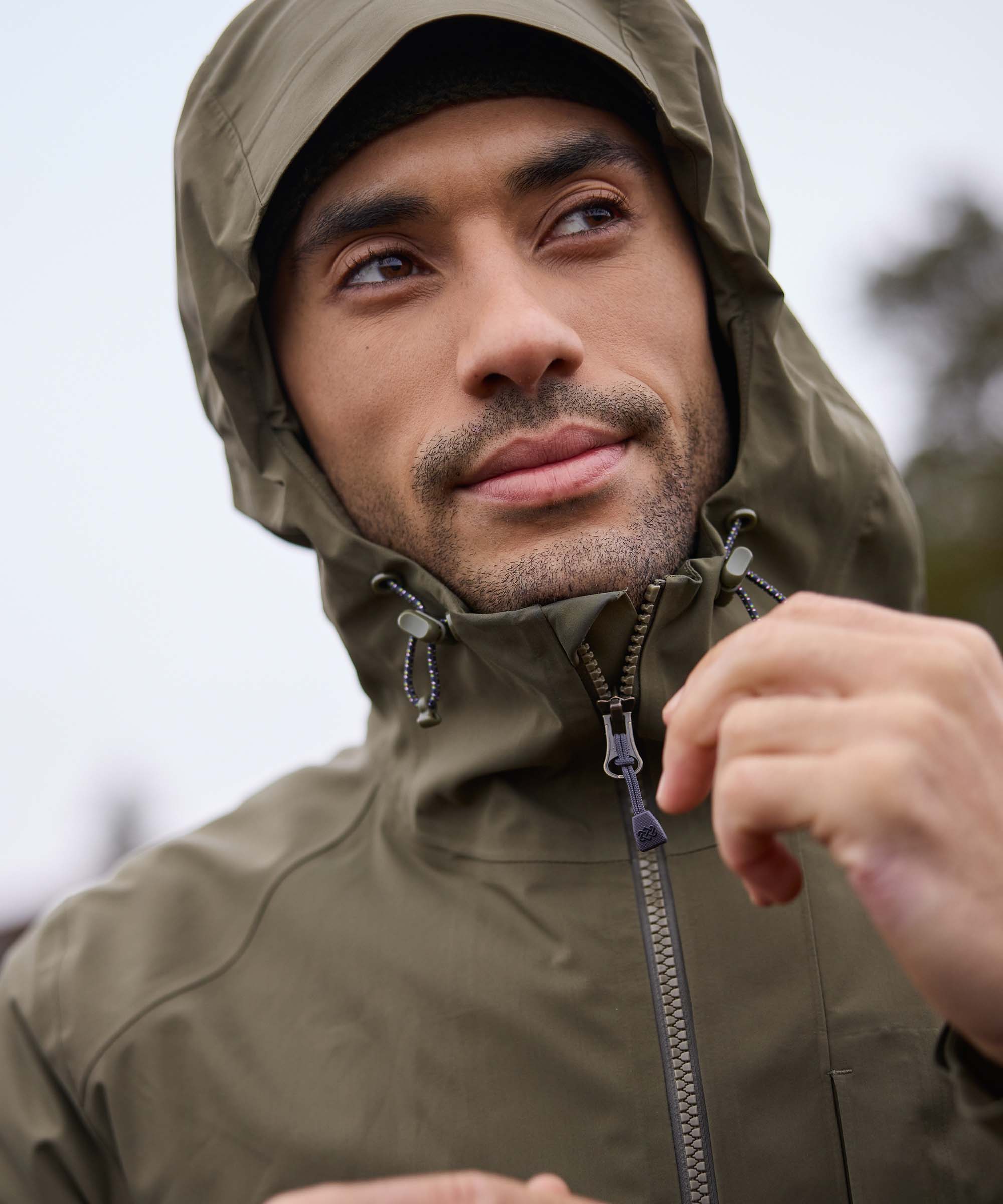 A close up of a man wearing a green hooded waterproof jacket in a forested landscape. He is looking into the distance whilst zipping up the jacket and adjusting the hood.