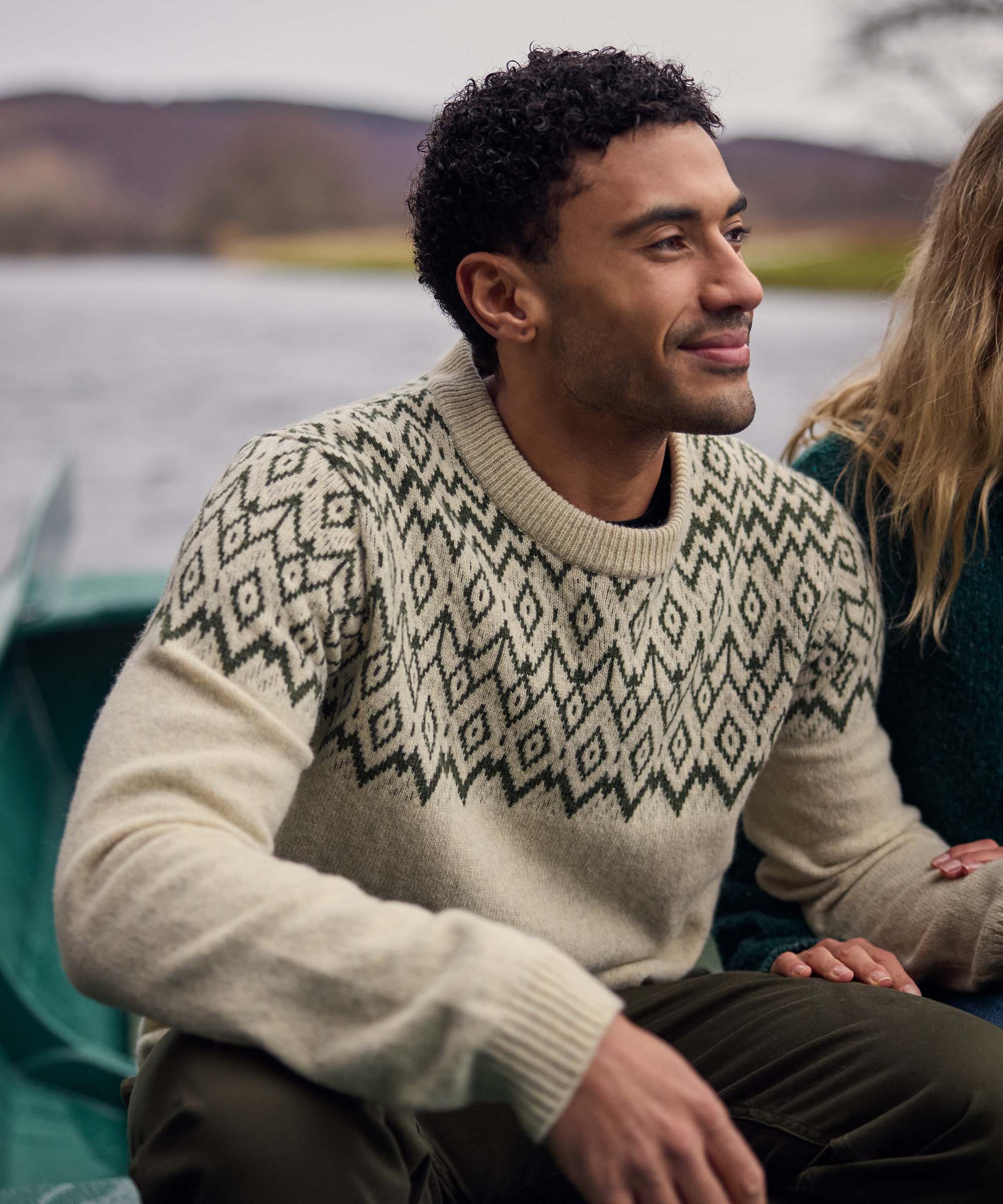 A man sits beside a lakeside campsite, smiling as he chats with someone nearby. He wears a cream crew-neck knit sweater featuring a bold black geometric pattern across the shoulders and chest. The relaxed outdoor setting and casual posture create a warm, social atmosphere.