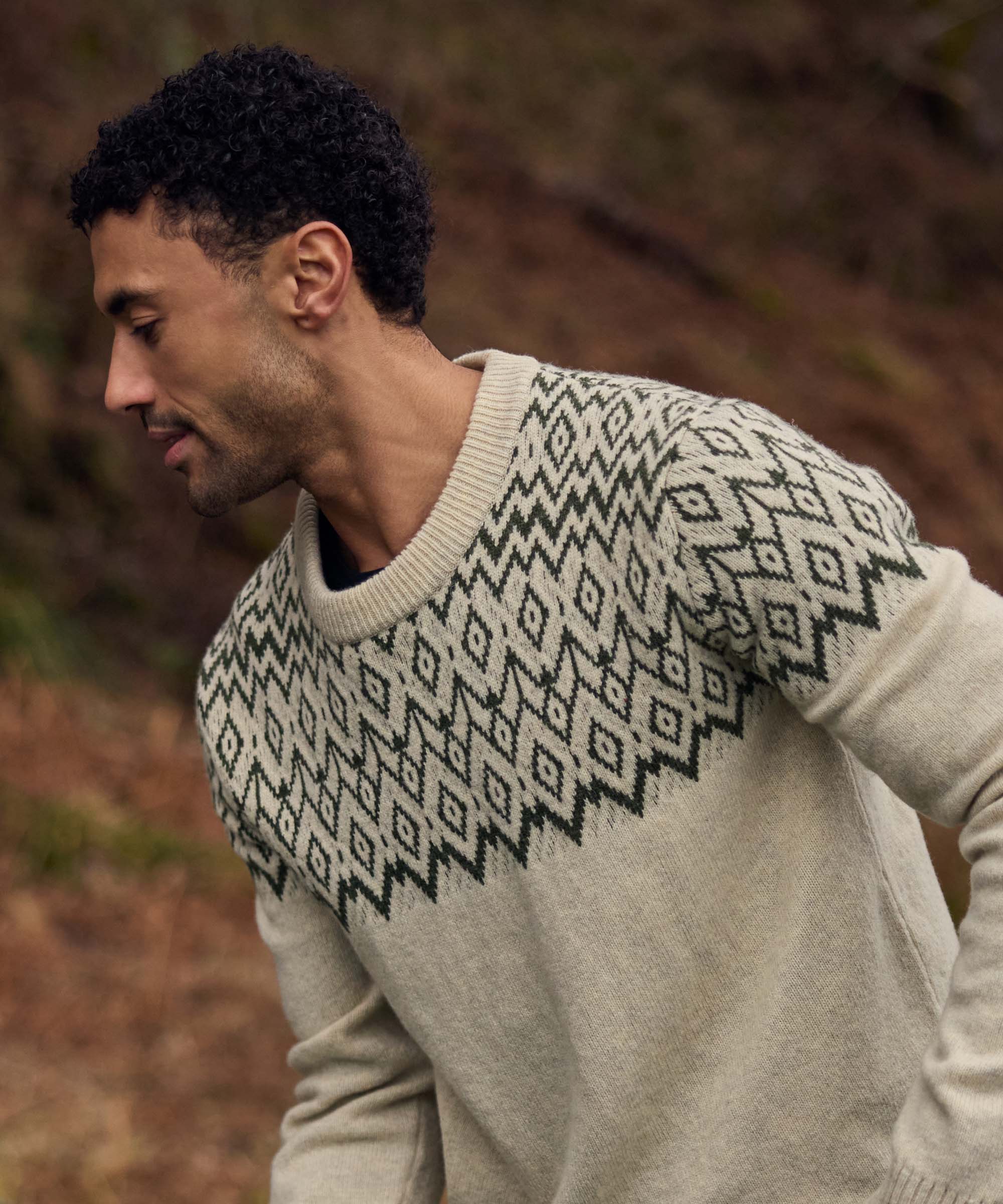 A man moves through a rugged outdoor landscape, leaning slightly forward as if stepping over uneven ground. He wears a cream knit crew-neck sweater with an intricate black geometric yoke pattern that extends across the shoulders and upper sleeves. The earthy tones of the surrounding hills complement the traditional-inspired knit design.