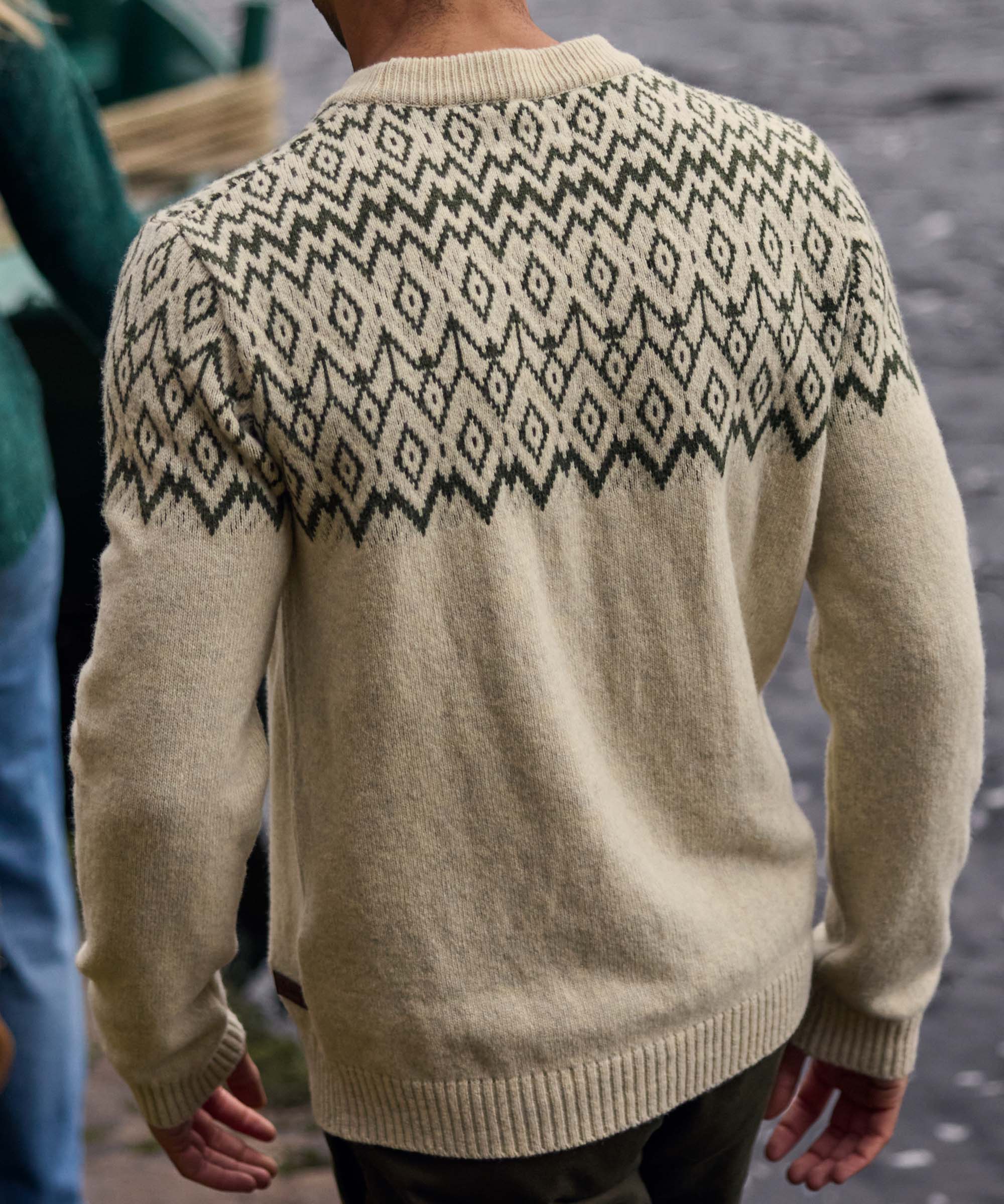 Rear view of a man walking outdoors near a lake, showcasing the back of a cream knit sweater with an all-over black geometric pattern across the shoulders and upper back. The detailed knit motif wraps around the garment, while the natural waterside setting provides a scenic backdrop.
