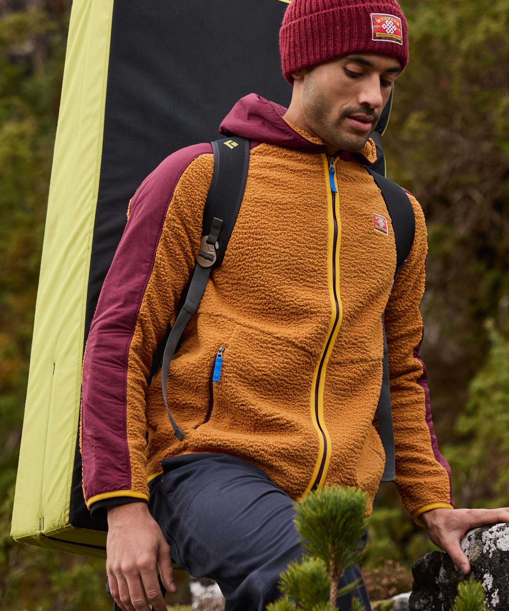 A man stands beside a tent in a rocky campsite, looking down as he navigates uneven terrain. He wears a hooded fleece jacket with a textured golden-brown body, burgundy sleeves and hood, bright yellow zip detailing, and a backpack. The image captures a moment of movement during an outdoor camping adventure.