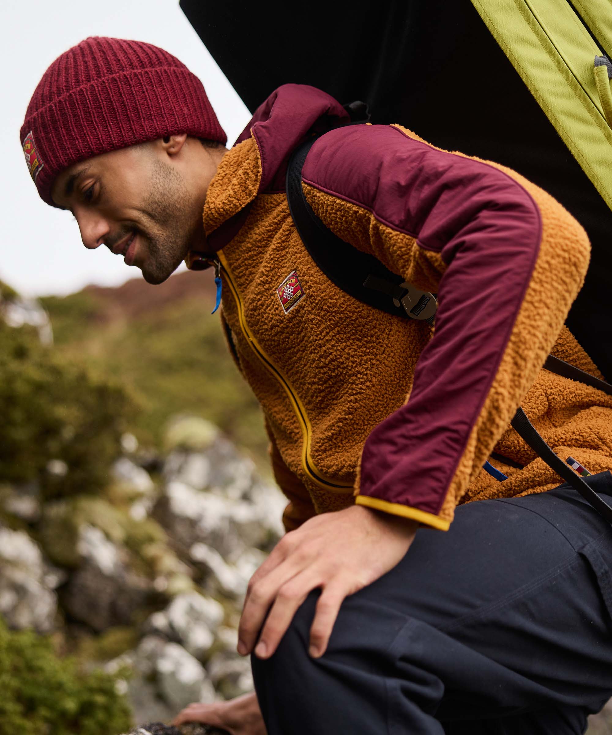 A man carefully lowers himself onto a rock near a tent, smiling as he settles into place. He wears a colour-block hooded fleece jacket with a golden-brown fleece body, burgundy sleeves, and contrasting yellow trim, paired with a burgundy knit beanie. The campsite and rocky landscape create an authentic outdoor scene.