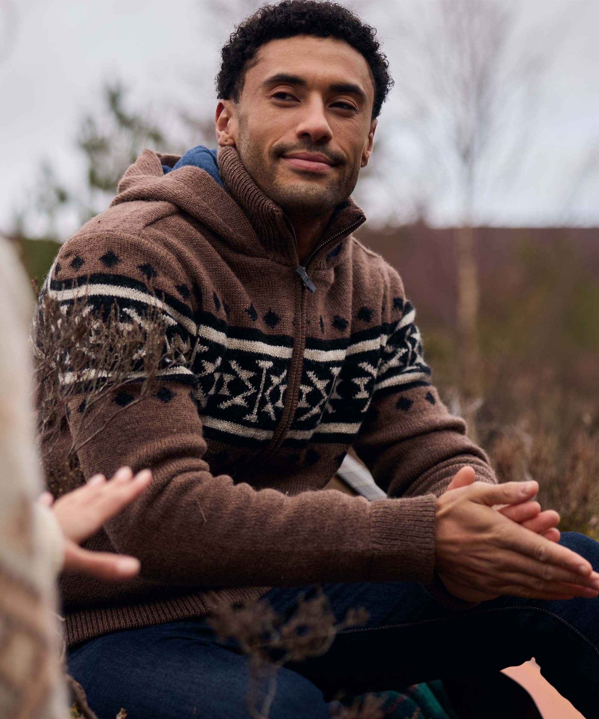 A man sits outdoors with a group beside a lake, listening attentively during a conversation. He wears a brown hooded knit sweater featuring bold black-and-cream Nordic-inspired patterns across the chest and sleeves. The relaxed gathering and natural surroundings create a warm, communal atmosphere.