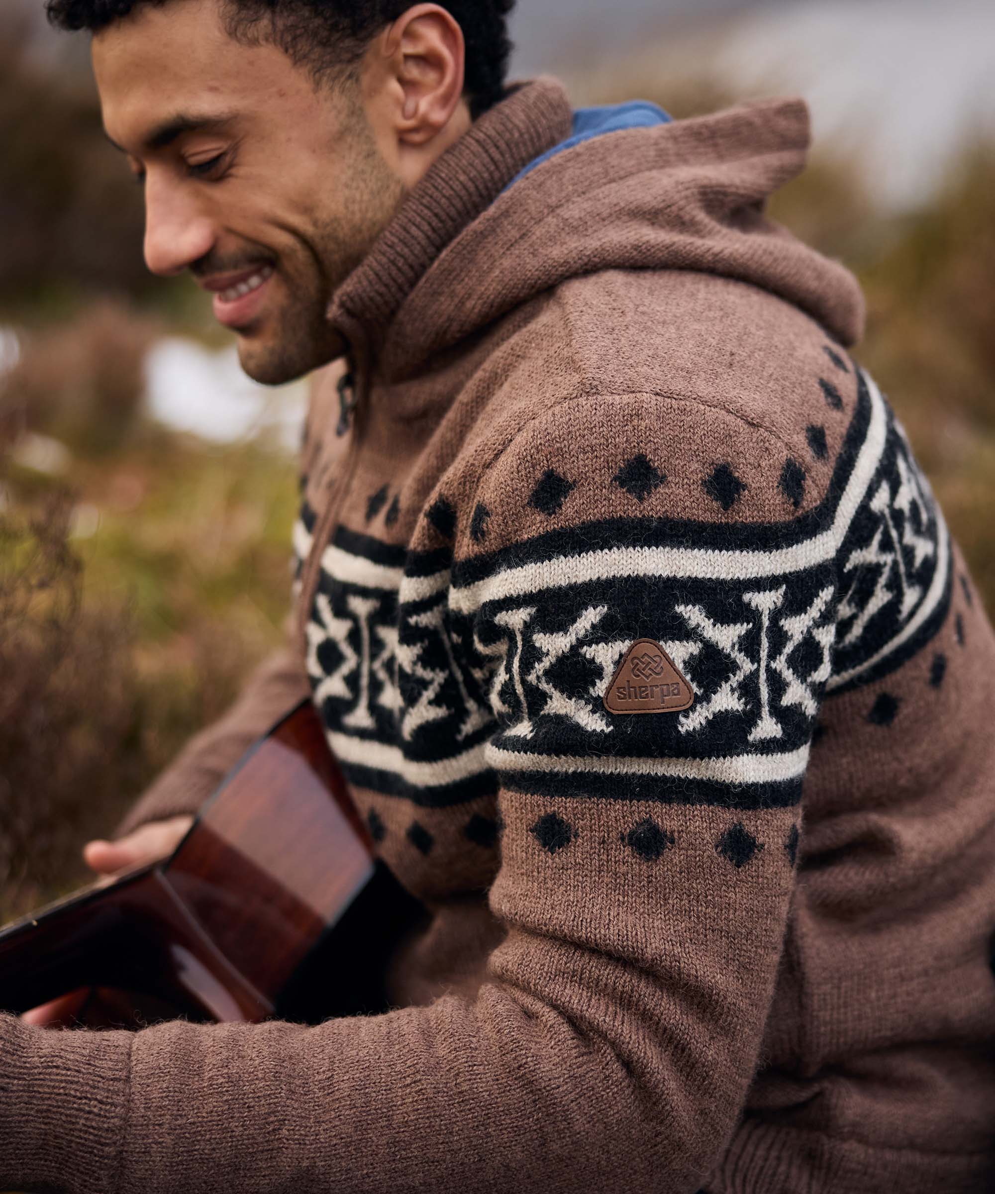 A man sits outdoors in a rugged countryside setting, smiling as he holds an acoustic guitar in his lap. He wears a brown hooded knit sweater featuring bold black-and-cream Nordic-inspired patterns across the chest and sleeves. The candid moment suggests a relaxed gathering in nature, with blurred vegetation and earthy tones in the background.