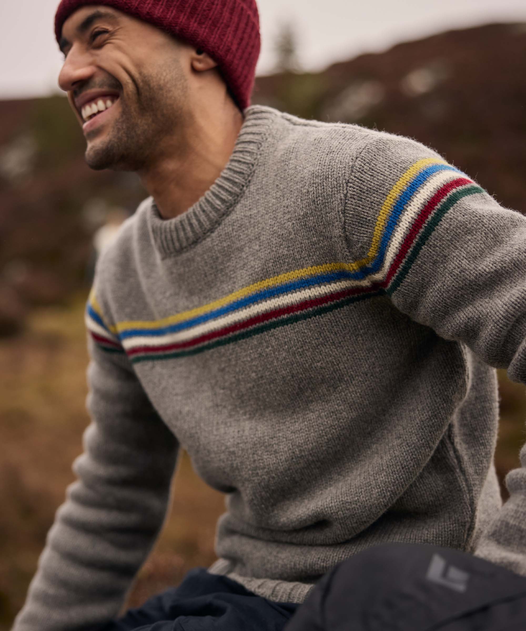 A man wearing a burgundy knit beanie laughs while seated outdoors on a hillside. He is dressed in a grey crew-neck sweater with colourful horizontal stripes across the chest, creating a vibrant contrast against the muted landscape. The image captures a cheerful moment shared during an outdoor adventure.