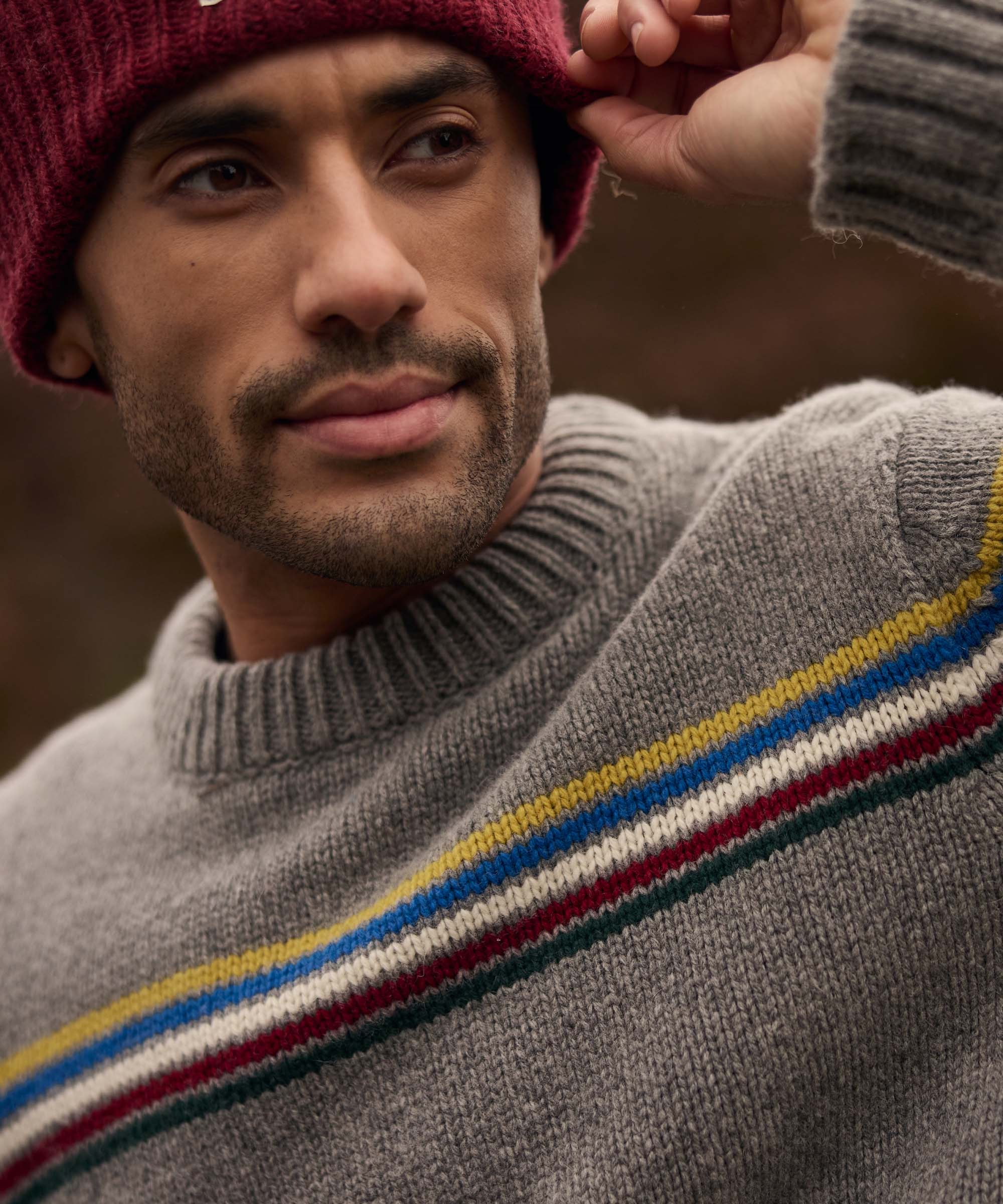 Close-up portrait of a man adjusting the edge of his burgundy knit beanie while looking off into the distance. He wears a grey crew-neck sweater with multicoloured stripe detailing across the chest and shoulders. The blurred mountain landscape behind him creates a warm and relaxed outdoor atmosphere.