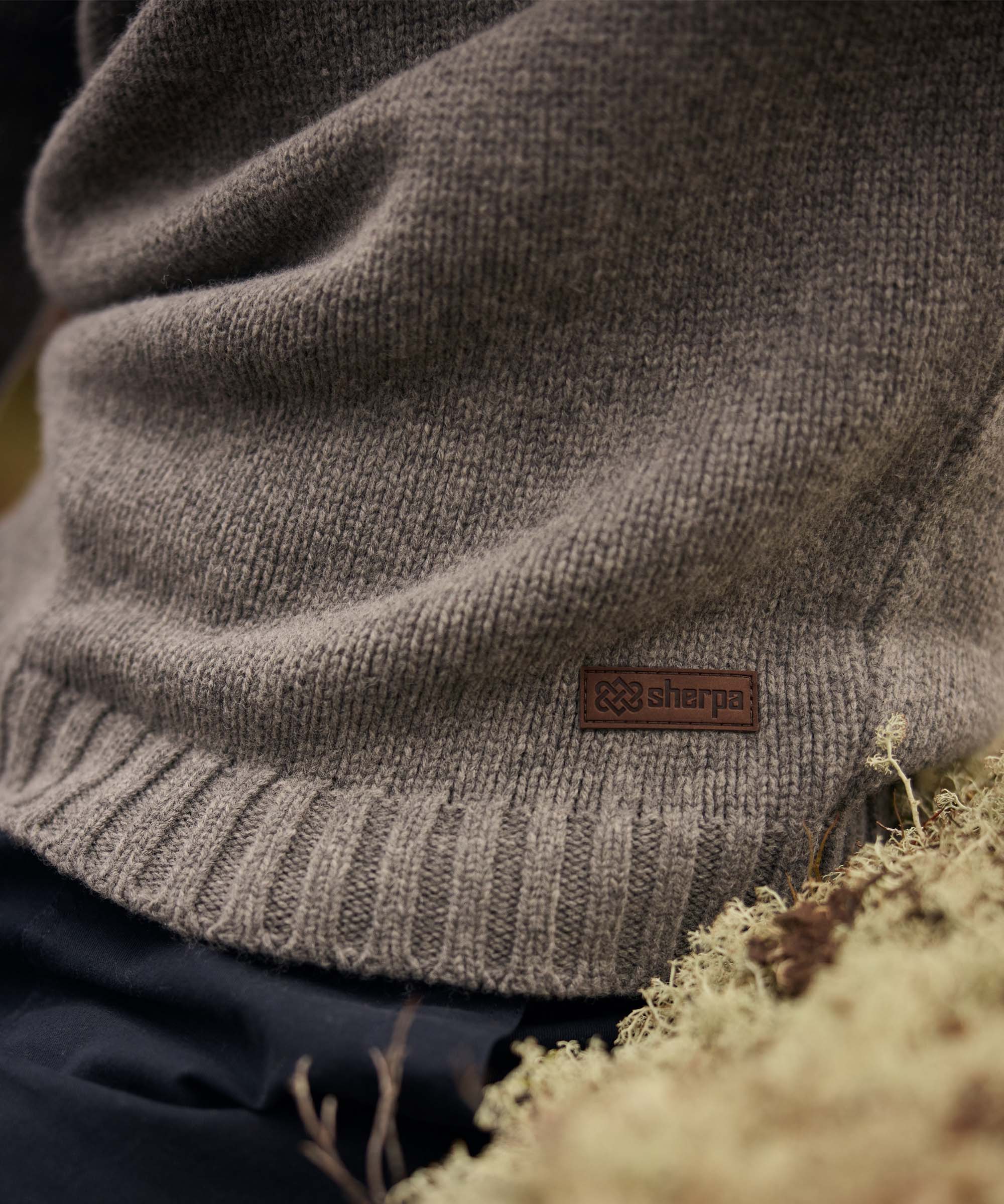 Detailed close-up of the lower portion of a grey knit sweater, highlighting the ribbed hem and subtle leather logo patch near the waist. The wearer is seated outdoors among natural vegetation, with soft-focus grasses and earthy textures framing the garment.