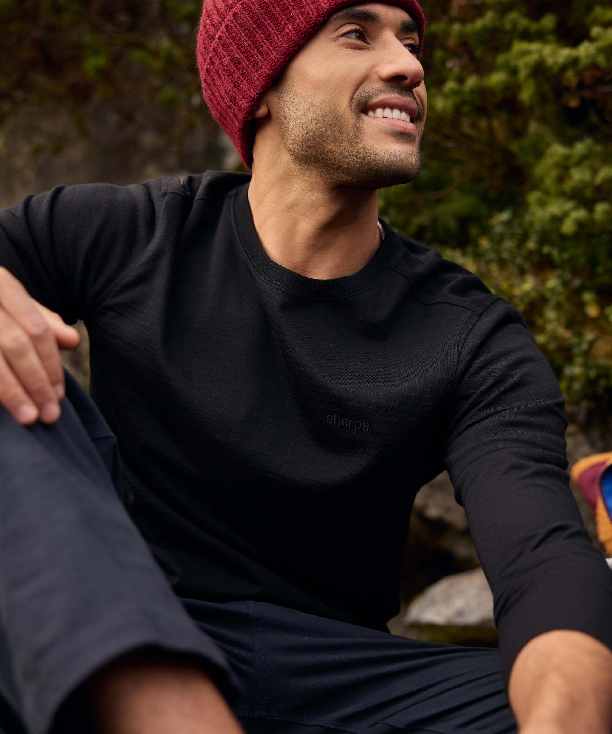 A man sits on rocky ground surrounded by greenery, smiling as he looks upward toward someone nearby. He wears a fitted black long-sleeve crew-neck shirt and a burgundy knit beanie, creating a simple, understated outdoor look. The image captures a relaxed social moment during a break in nature.