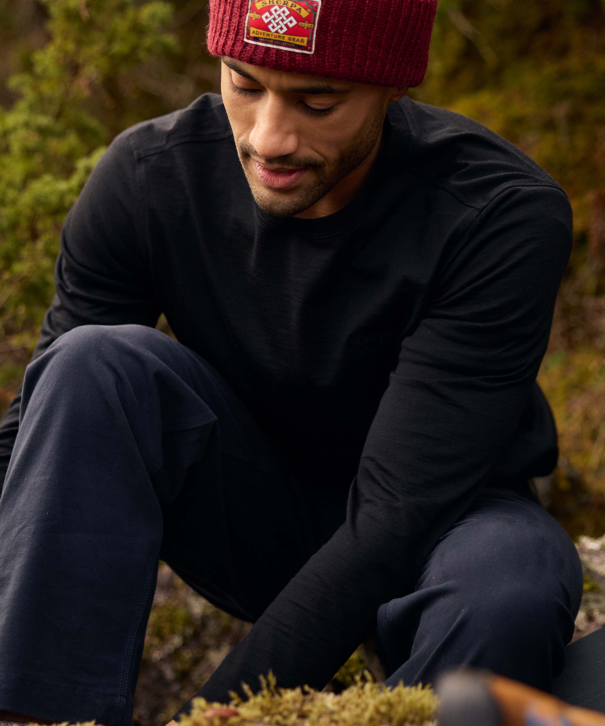 Seated on the ground in a natural landscape, a man looks down thoughtfully while resting his arms on his knees. He wears a black long-sleeve crew-neck shirt, dark trousers, and a burgundy beanie. The candid pose conveys a quiet pause during an outdoor excursion.