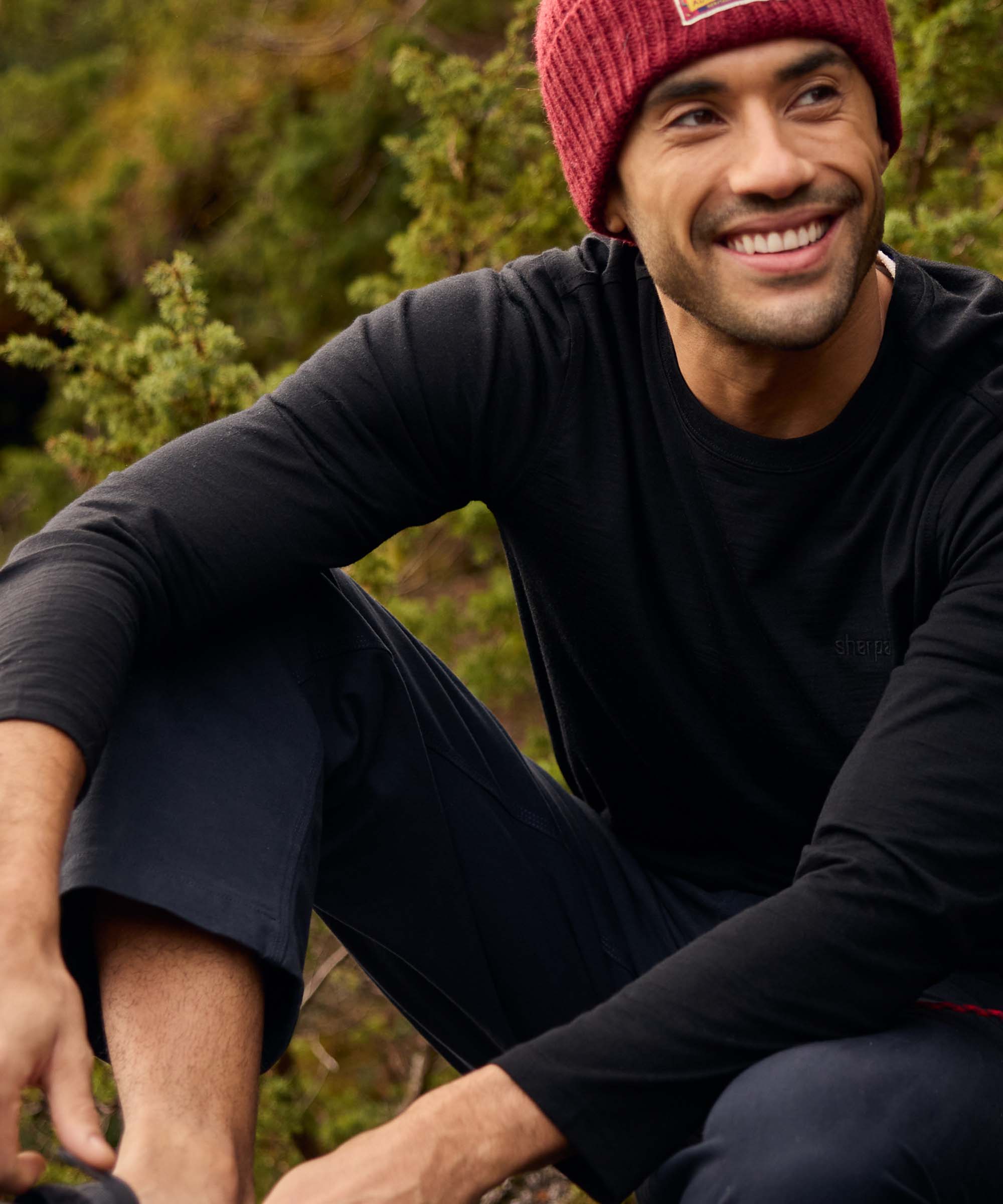 A smiling man relaxes outdoors among shrubs and rocks, seated comfortably with one arm resting on his knee. He wears a black long-sleeve crew-neck shirt paired with a burgundy knit beanie. The image conveys warmth, companionship, and enjoyment of the natural surroundings.