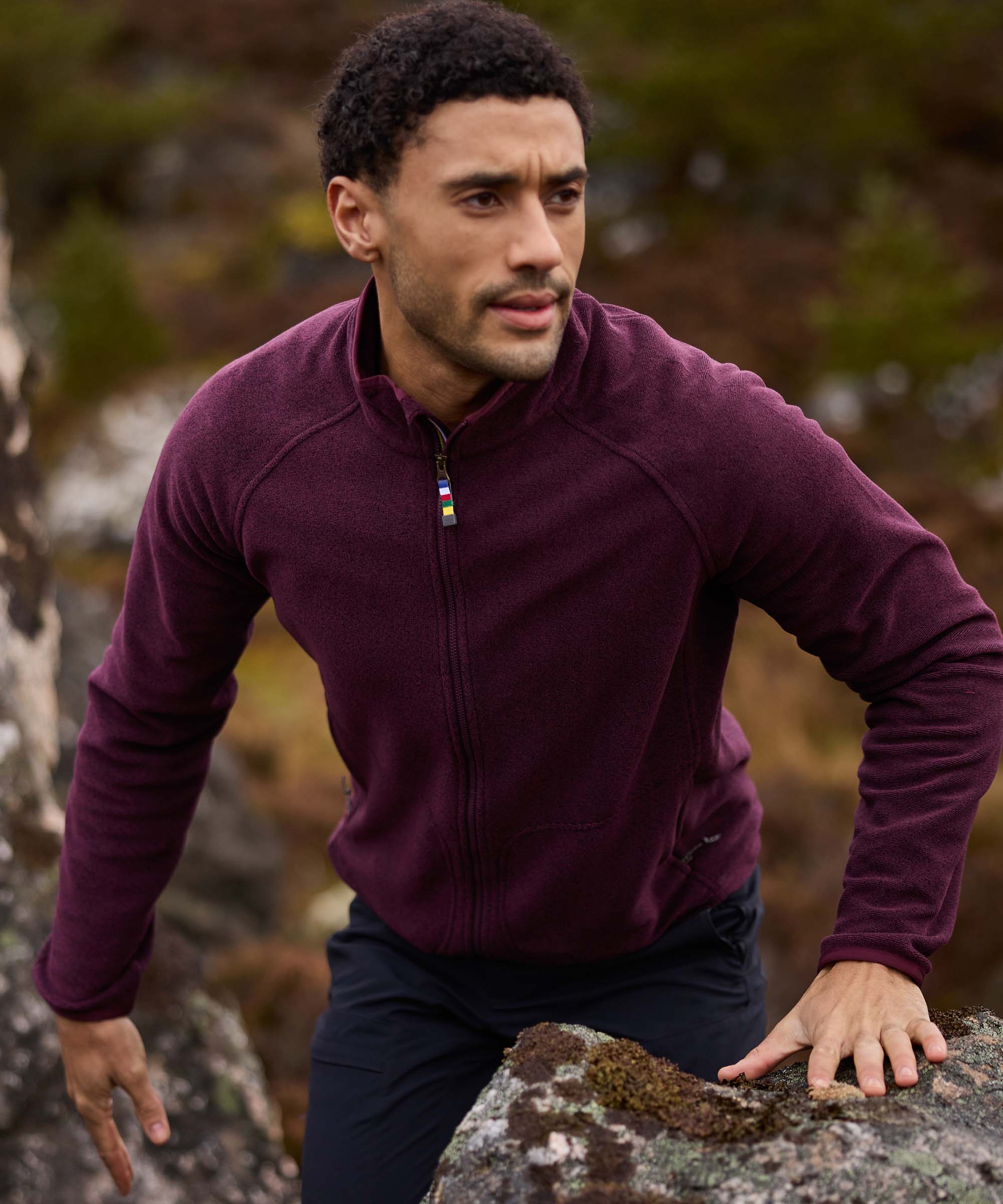 A man climbs over rocky terrain in a rugged outdoor setting, using his hands for balance as he moves uphill. He wears a burgundy full-zip fleece jacket with a high collar and contrasting trim at the neckline, paired with dark trousers. The image captures an active moment of exploration against a backdrop of rocks and moorland vegetation.