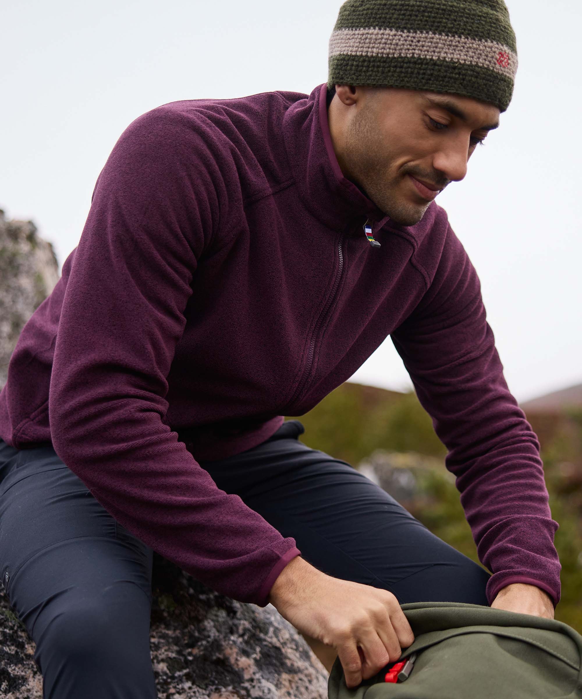 A mid shot of a man in a purple fleece jacket and green beanie sitting on a rock outdoors. He is looking in his backpack looking for his equipment.