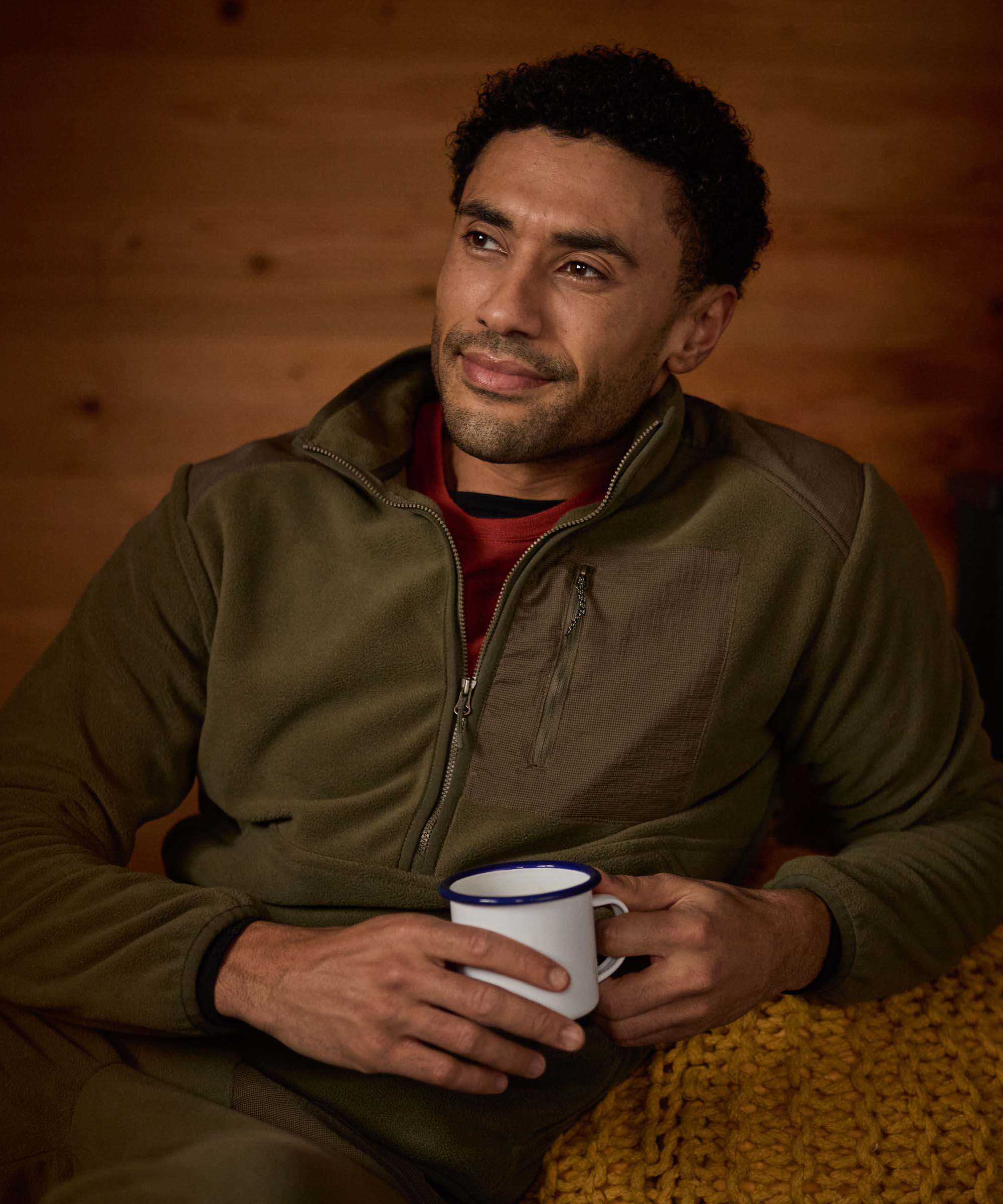 A man relaxes in a rustic wooden cabin, leaning back with one arm and holding onto a camping mug with his hand. He wears an olive-green half-zip fleece and a red shirt underneath.