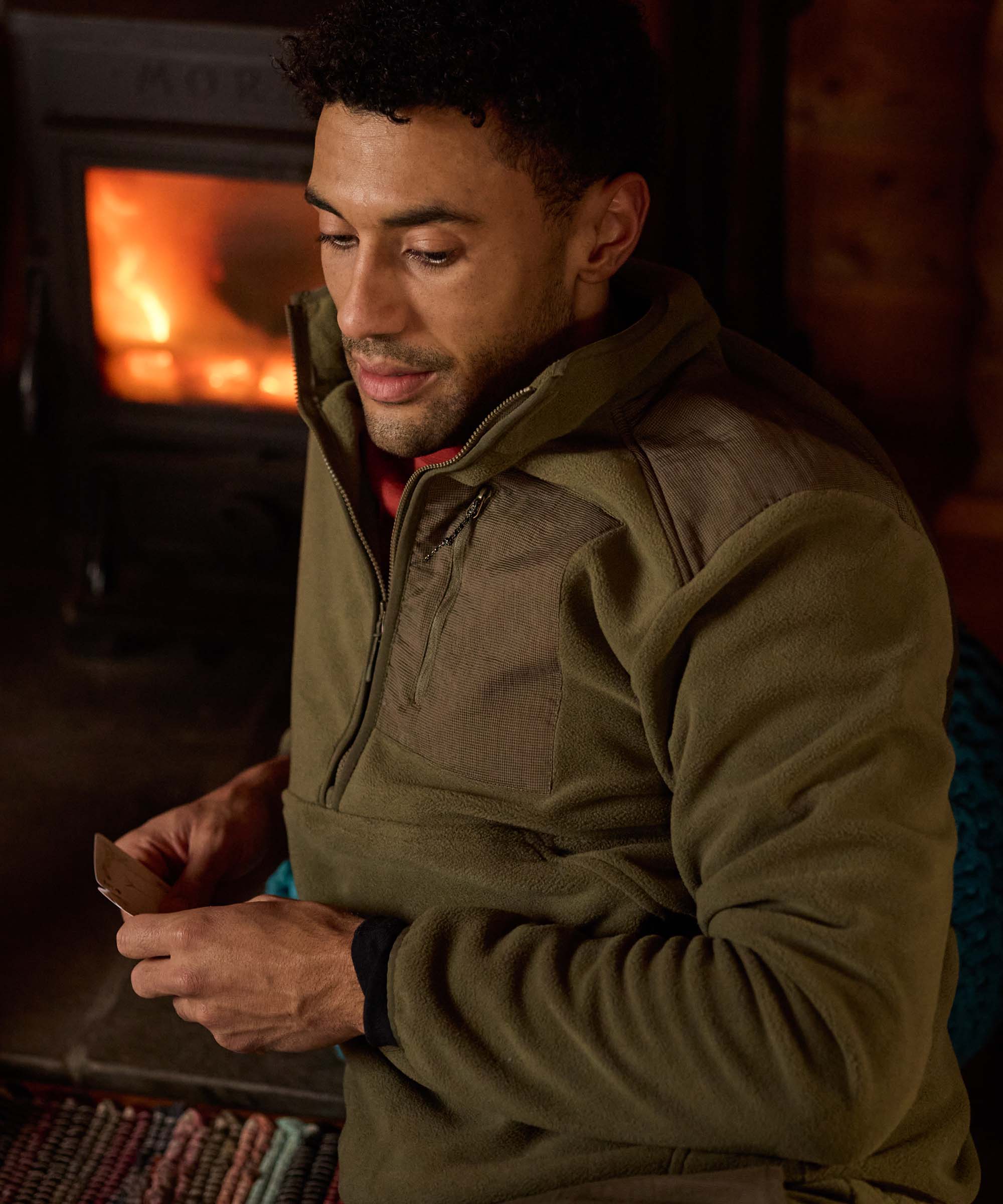 A man relaxing in a rustic wooden cabin near a wood burning stove. He is hold playing cards in his hands and is wearing  a olive-green half-zip fleece and a red shirt underneath.