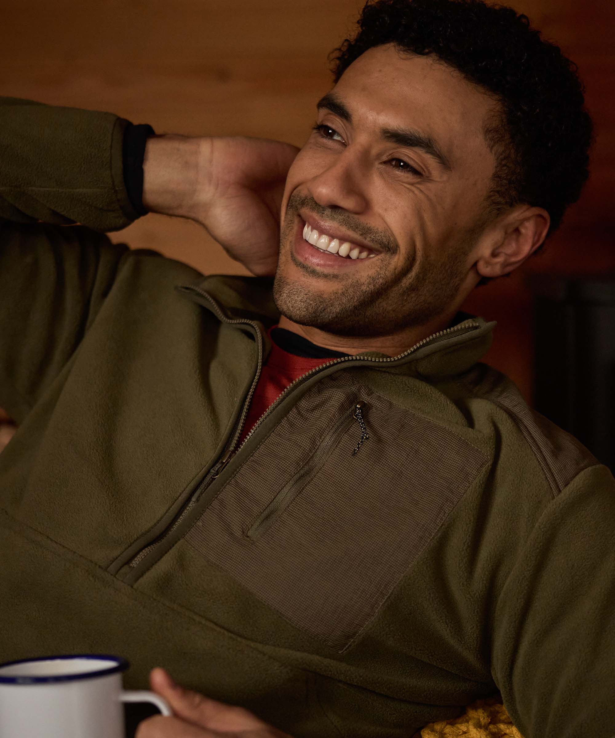 A man relaxes indoors in a rustic wooden cabin, leaning back with one arm behind his head and smiling warmly. He wears an olive-green half-zip fleece with a contrasting chest pocket and a red shirt underneath. A mug sits nearby, creating a cozy aprs-adventure atmosphere.
