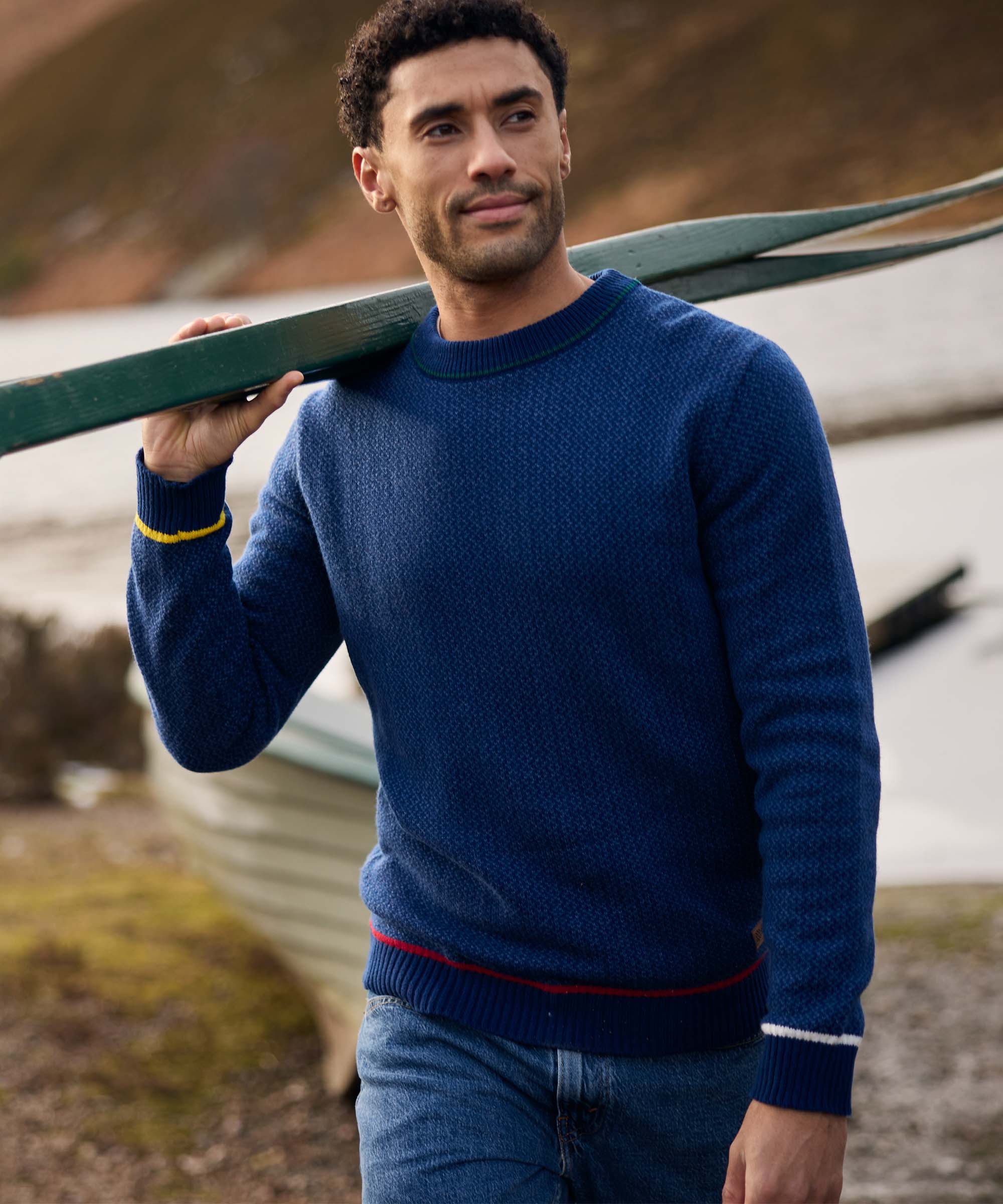 A man walks along a lakeside carrying a green canoe across his shoulder. He wears a textured blue crew-neck sweater with contrasting trim at the collar, cuffs, and hem, paired with jeans. Calm water and rolling hills form the scenic outdoor backdrop.