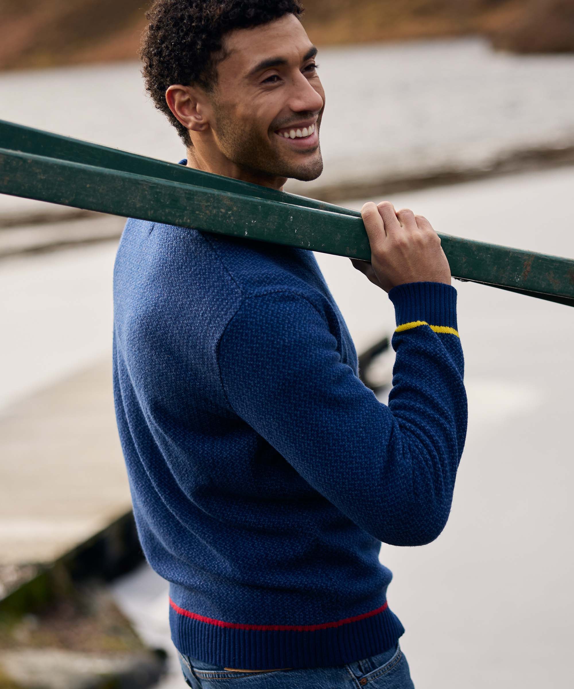 Smiling over his shoulder, a man carries a canoe while walking beside a lake. He is dressed in a blue knit crew-neck sweater with colourful accent detailing at the cuffs and hem. The image captures a candid moment during an outdoor excursion.