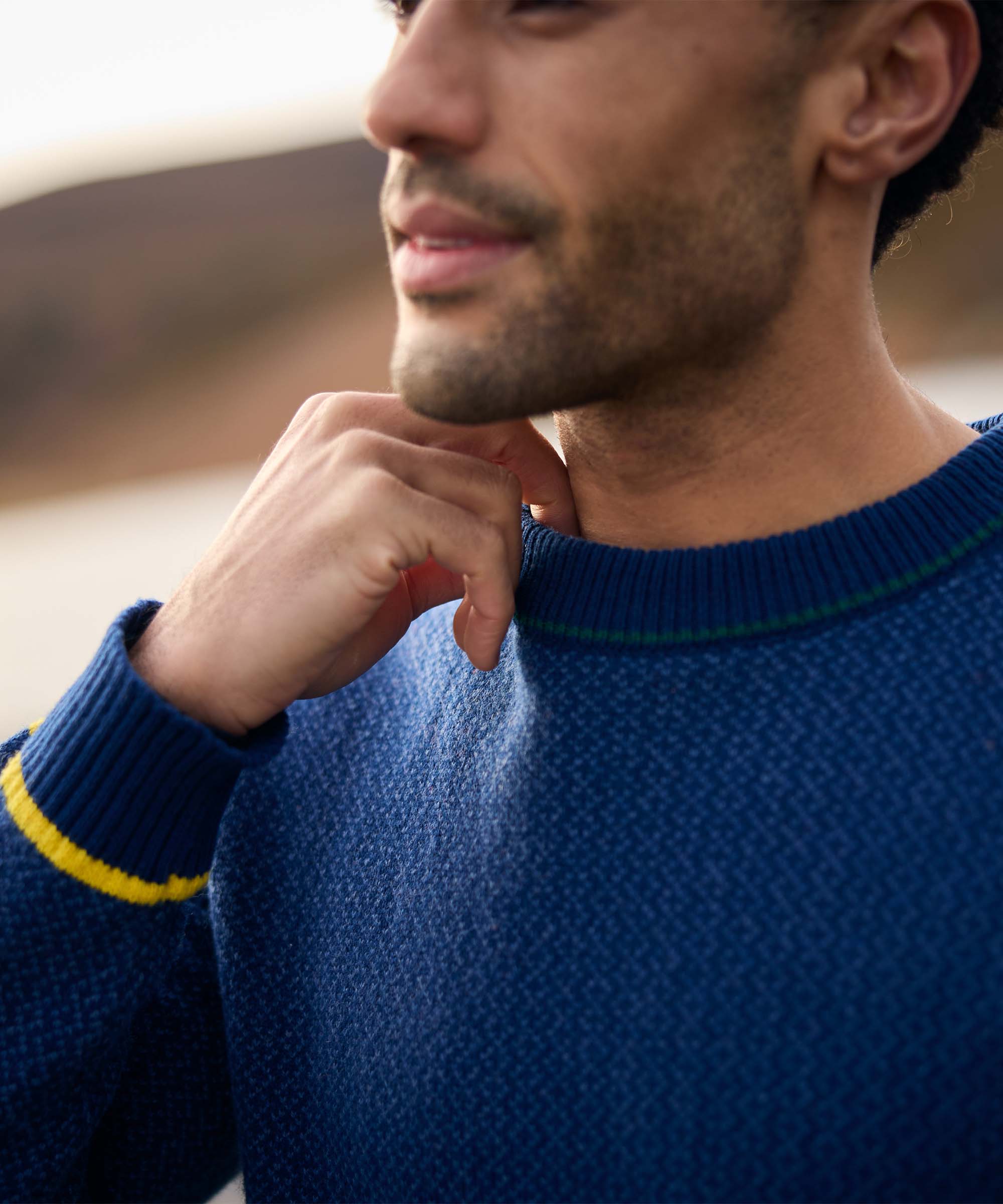 Close-up portrait of a man standing outdoors near a lakeshore, resting his hand against his chin. He wears a textured blue crew-neck sweater featuring subtle contrast trim at the neckline and cuffs. The shallow depth of field keeps focus on the sweater and relaxed expression.