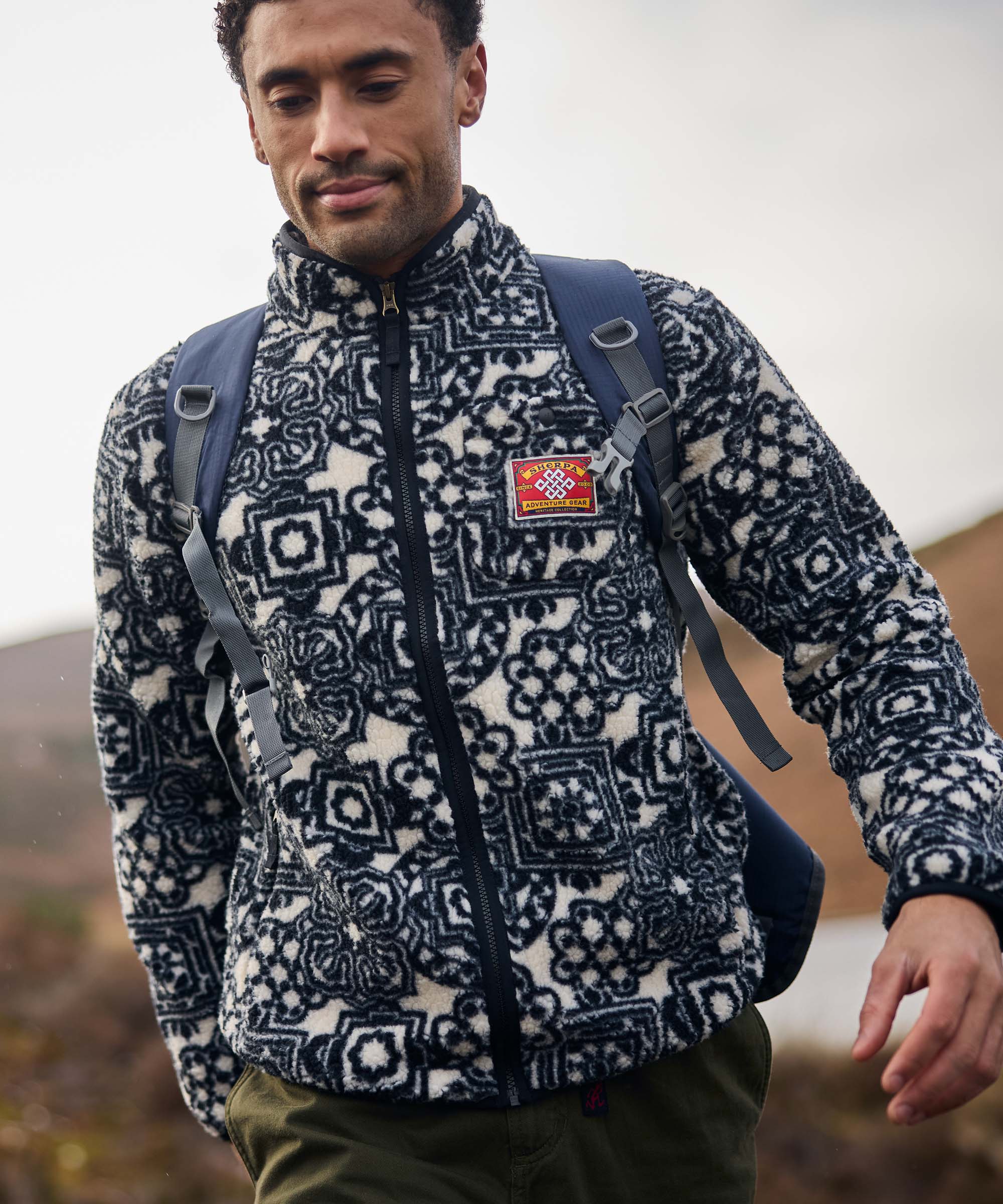 A man hikes through rugged countryside wearing a black-and-white printed full-zip fleece jacket and a backpack. He looks ahead while walking across uneven terrain, with hills and open landscape stretching behind him.