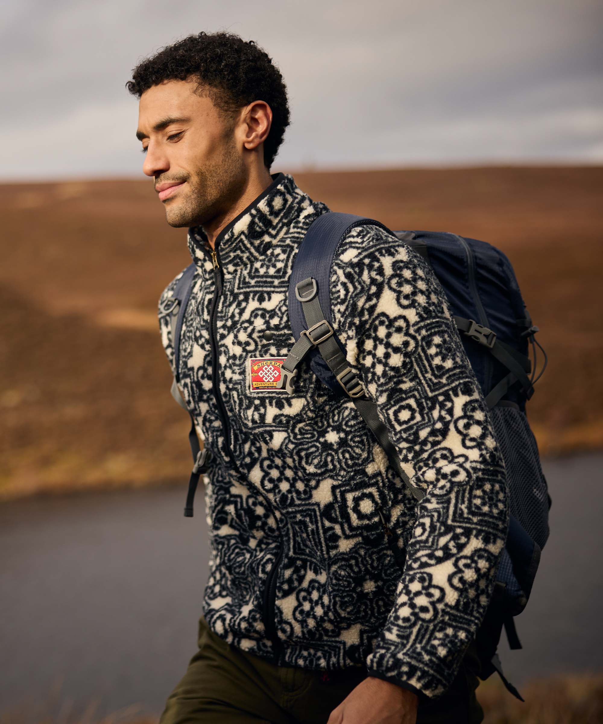 Walking beside a lake, a man carries a backpack and smiles softly while enjoying the scenery. He wears a patterned black-and-white fleece jacket with a full-length zip and high collar. The earthy landscape and water create a peaceful outdoor setting.