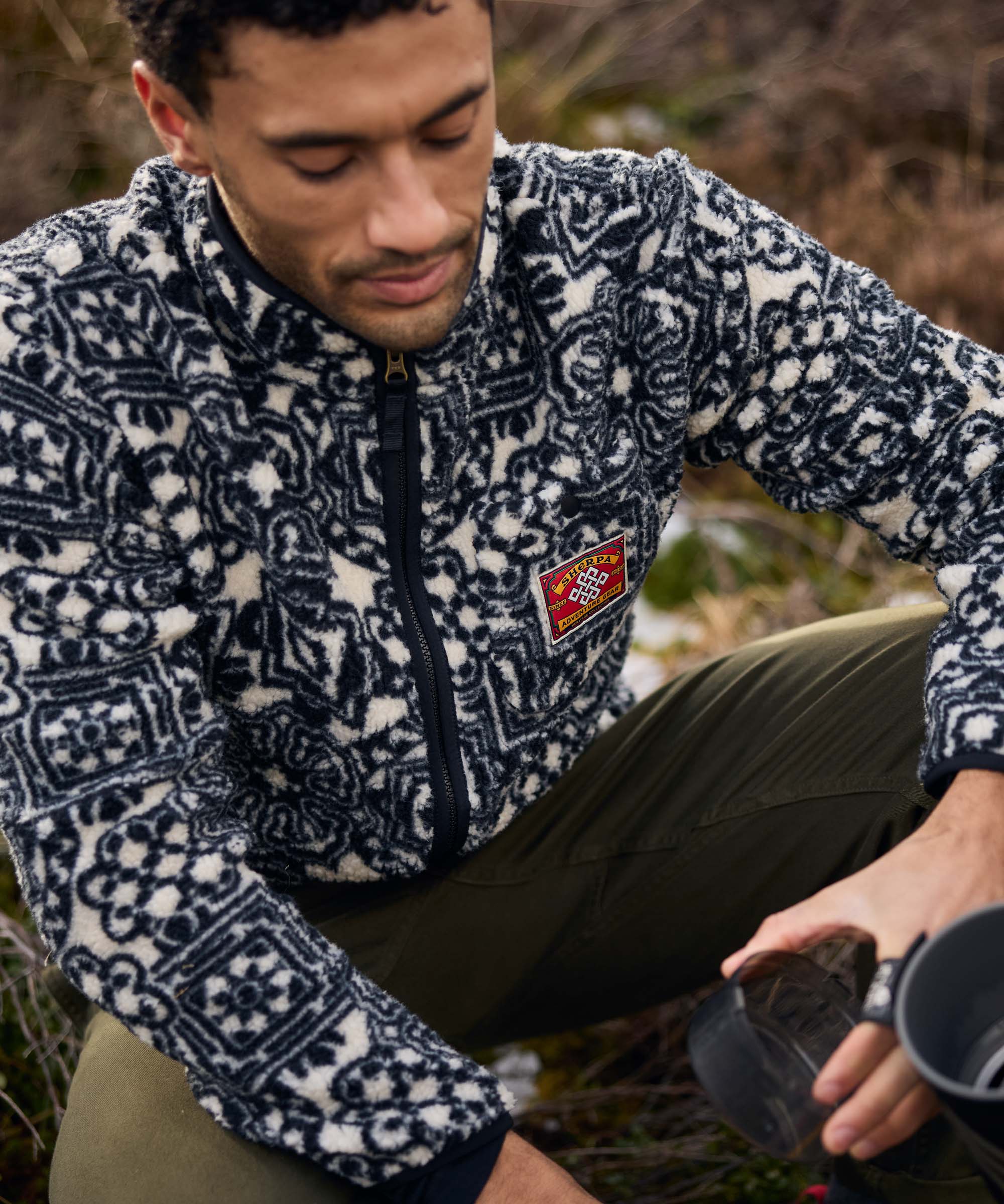 A man crouches on rocky ground while taking a break outdoors. He wears a black-and-white geometric print fleece jacket and olive trousers, looking down as he adjusts or reaches for gear beside him. The image conveys a quiet moment during a hike.