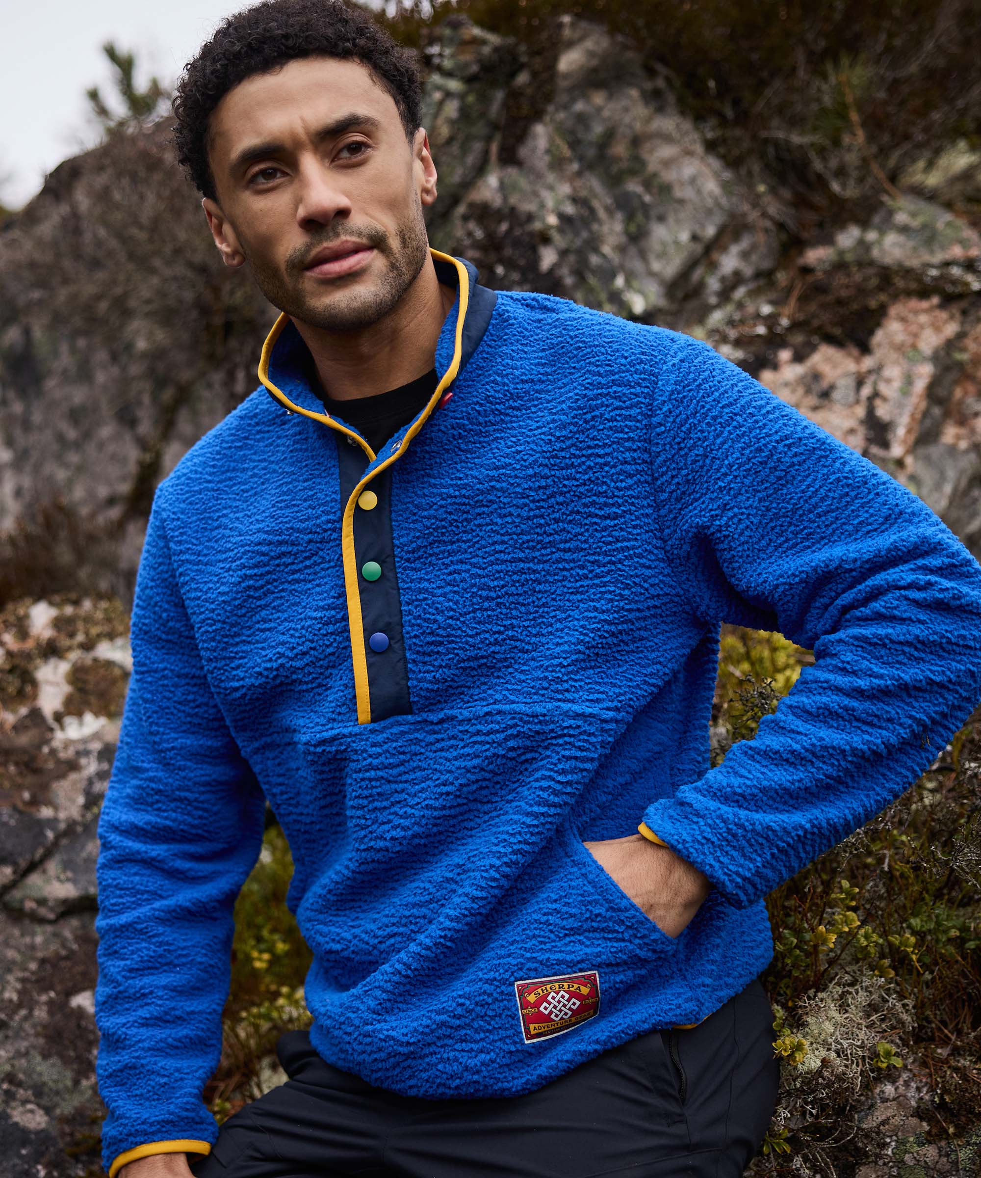 A man sits on rocks in a rugged natural landscape, posing casually with one hand in his pocket. He wears a bright blue quarter-snap fleece pullover with contrasting yellow trim around the placket and collar. The outdoor setting highlights a relaxed adventure-inspired lifestyle.