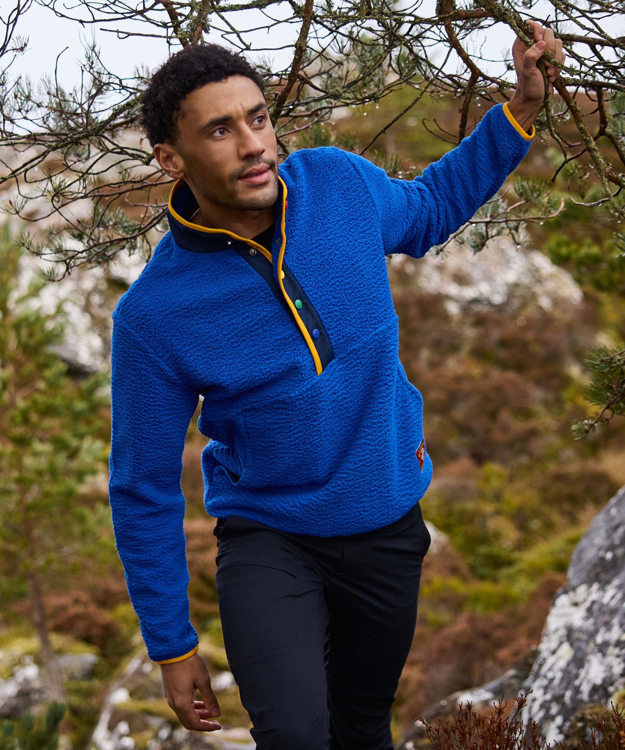 A mid shot of a man wearing a blue fleece pullover with yellow trim in a forest setting. The fleece is paired with black hiking trousers. The is hiking through the forested area moving branches out the way with his hands.