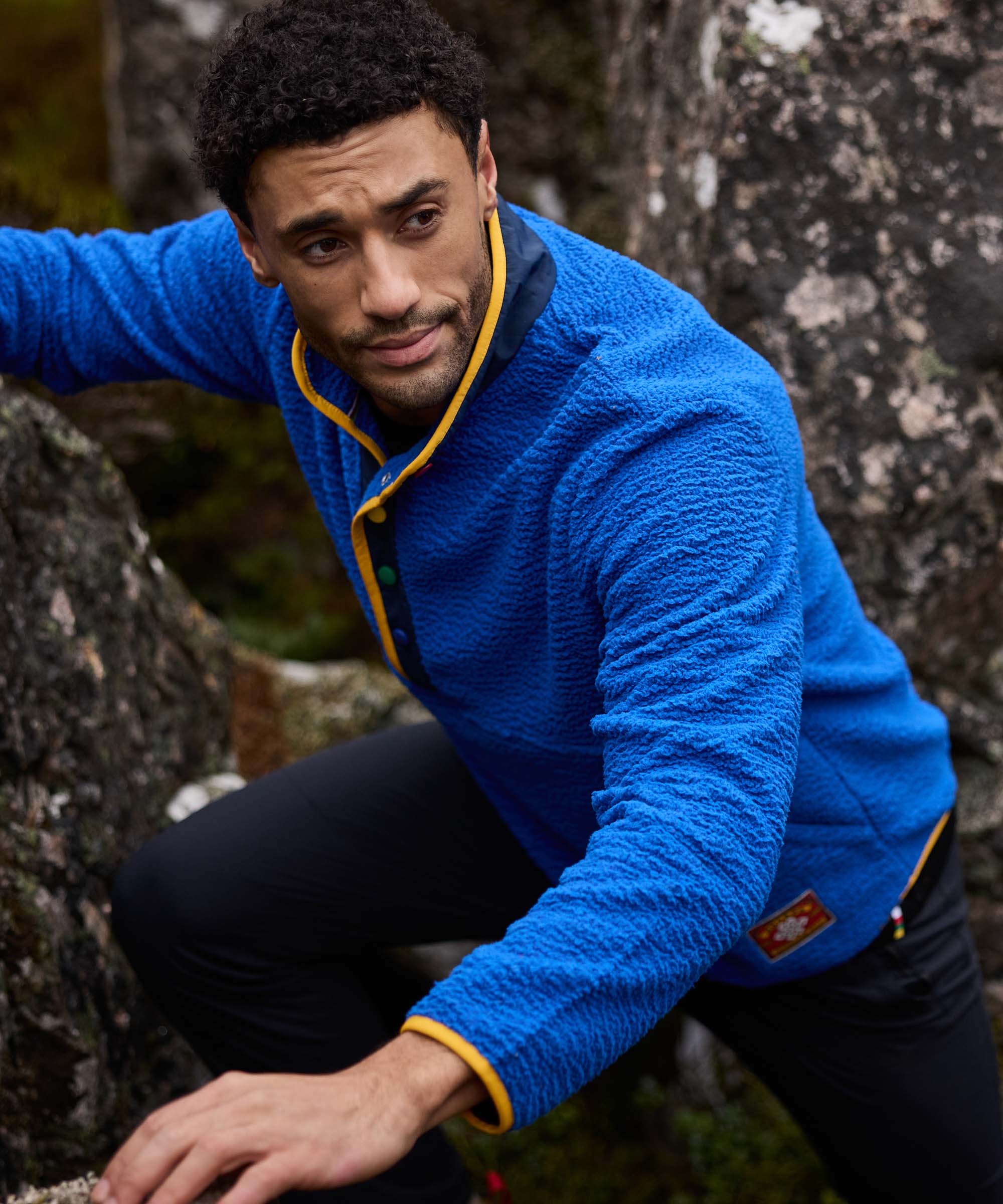 A mid shot of a man wearing a blue fleece jacket hiking in a rocky area. The fleece features yellow trim and colored pop button studs and a Sherpa Adventure Gear patch on the pocket. The man is climbing over large grey rocks whilst looking to one side.
