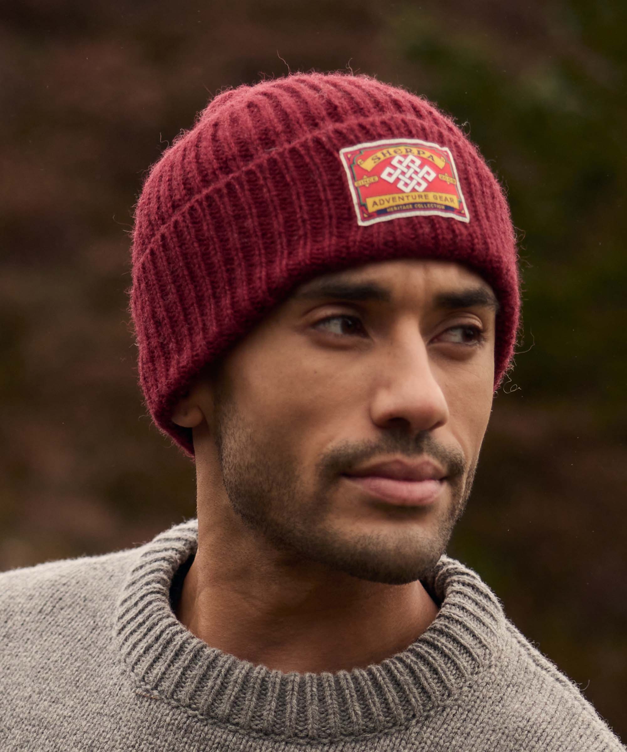 A man wearing a red knit beanie with a logo, looking to the side. The beanie is paired with a grey, knitted sweater with a ribbed collar.