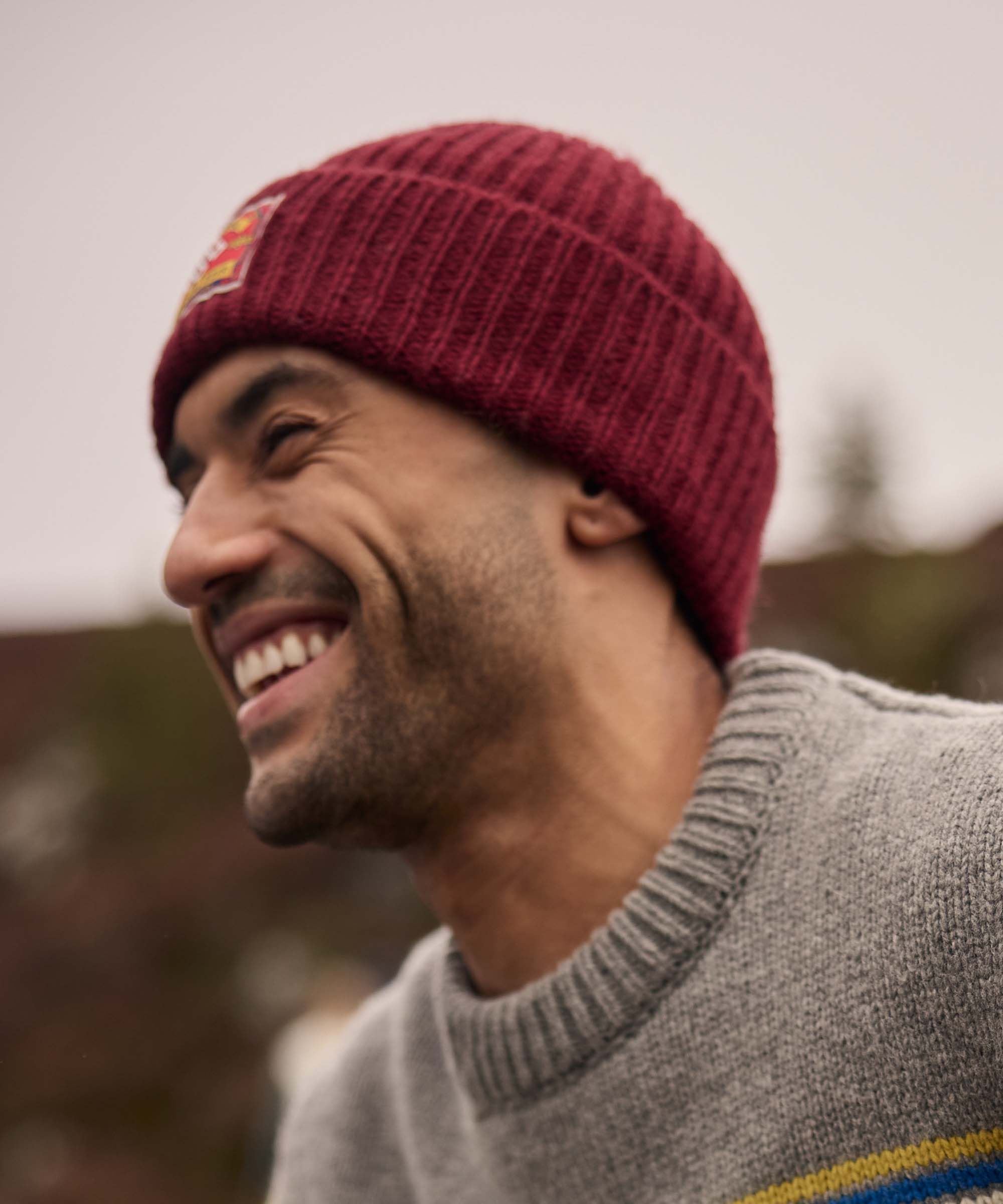 A man wearing a red knit beanie and grey sweater, smiling outdoors.