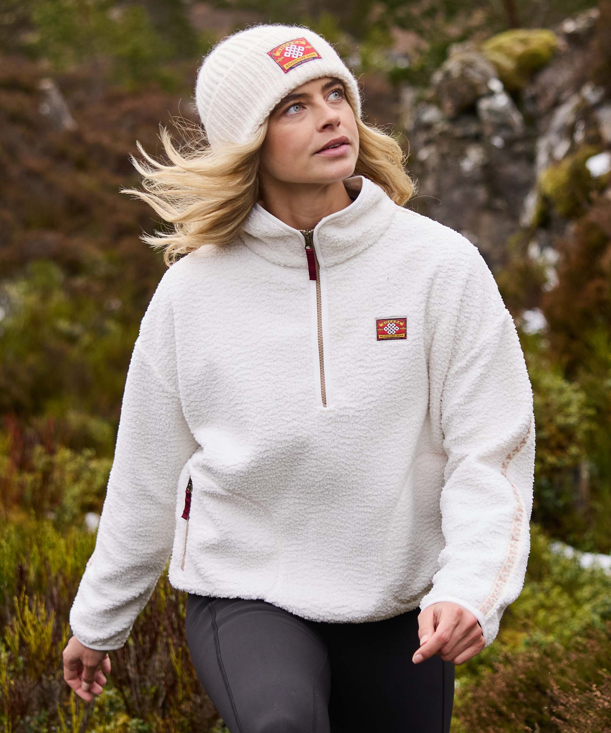 A woman walks through rocky moorland, looking upward as she explores the landscape. She wears a cream quarter-zip fleece pullover with a high collar, matching cream knit beanie, and dark leggings. The natural setting highlights a peaceful moment on the trail.
