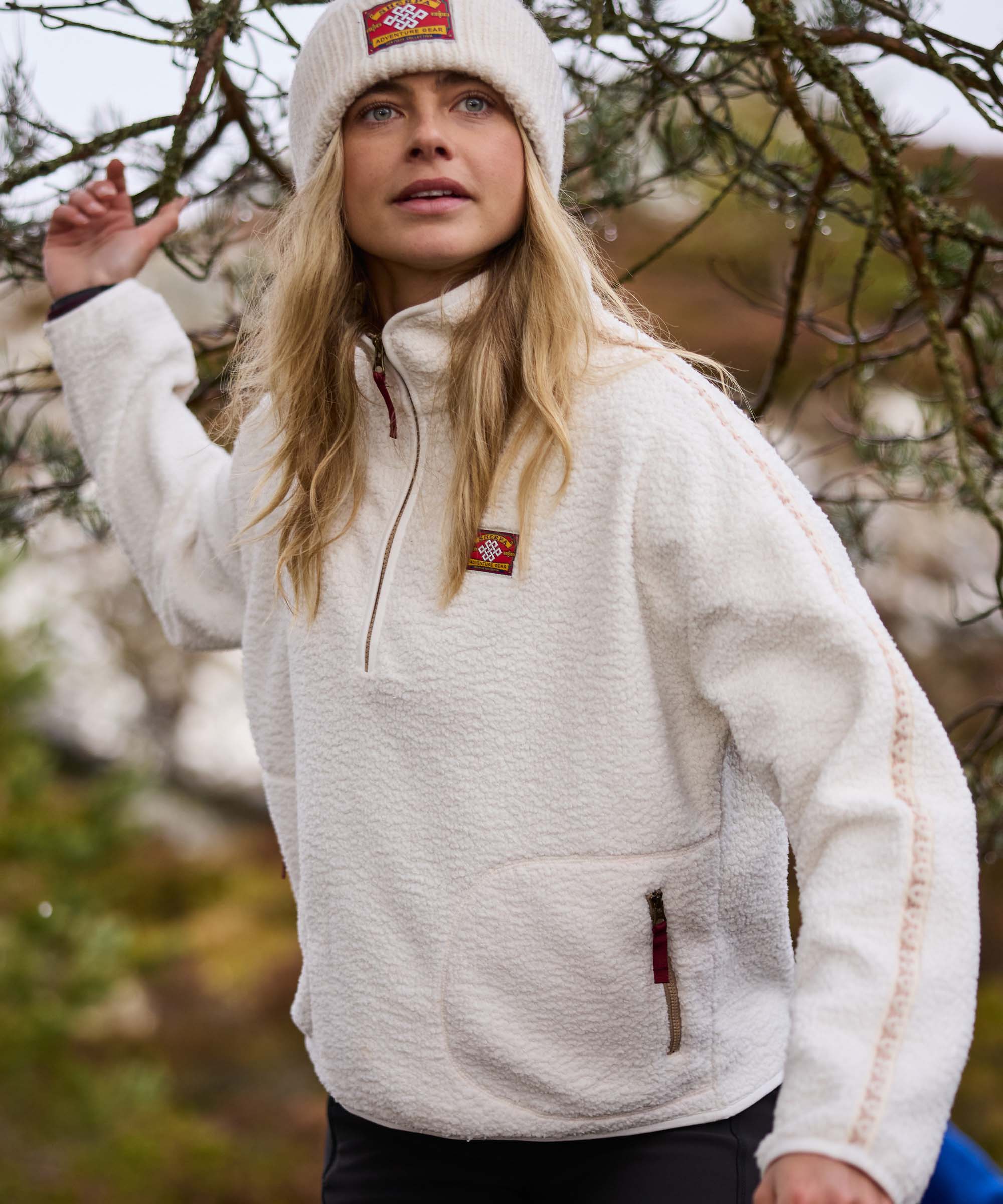 A woman reaches up to gently lift a low tree branch while walking through woodland. She wears a cream quarter-zip fleece pullover with a high collar and kangaroo pocket, paired with a cream knit beanie and dark leggings. The candid scene conveys curiosity and connection with nature.