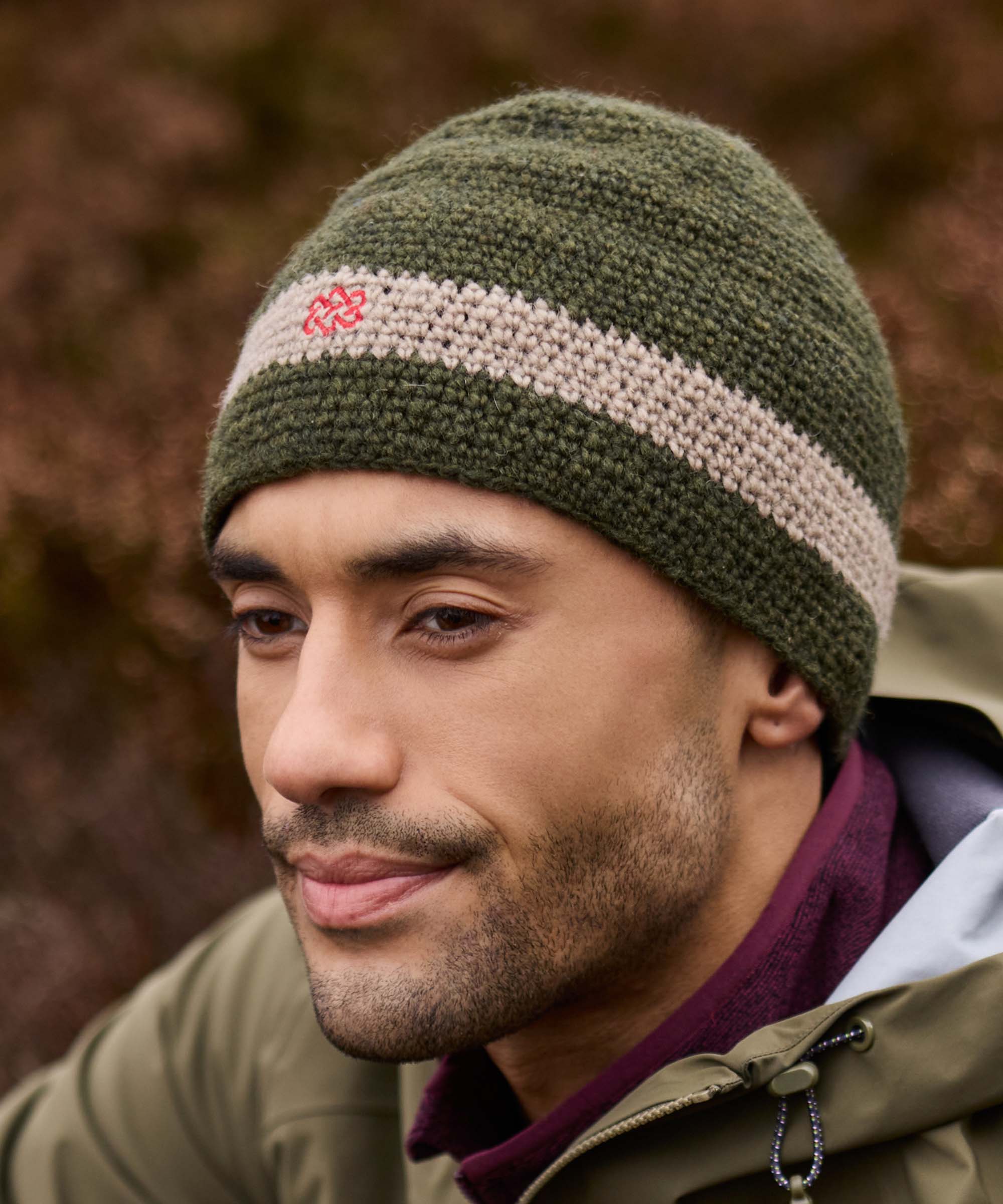 A close up of a man wearing a green knit beanie with a red logo. The hat is paired with a hooded, waterproof jacket and a red fleece. The man is hiking in the Scottish highlands with heather visible in the background of the image.