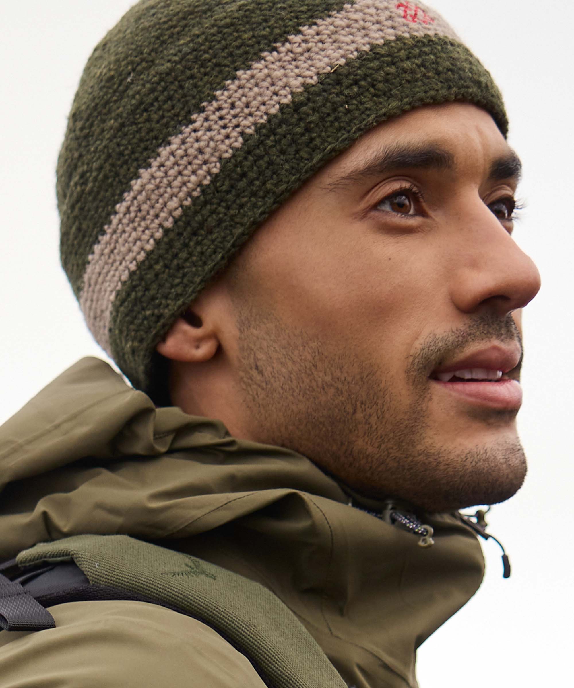 Close-up side profile of a person standing outdoors and looking into the distance, surrounded by a rugged natural landscape. The expression is calm and focused, as if taking in the view ahead. They wear an olive-green knit beanie with a contrasting cream stripe and an olive outdoor jacket.
