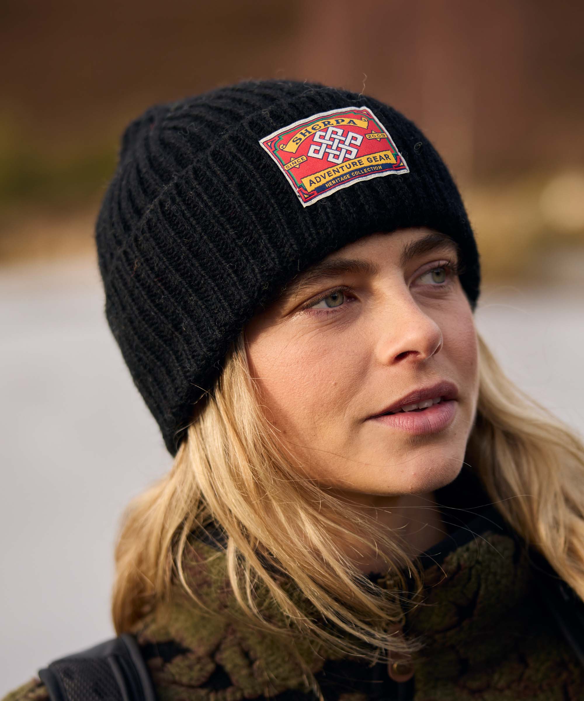 Close-up portrait of a woman outdoors, gazing thoughtfully into the distance. The background is softly blurred, emphasizing her expression and the natural surroundings. She wears a black rib-knit beanie with a woven patch and a dark patterned fleece jacket.