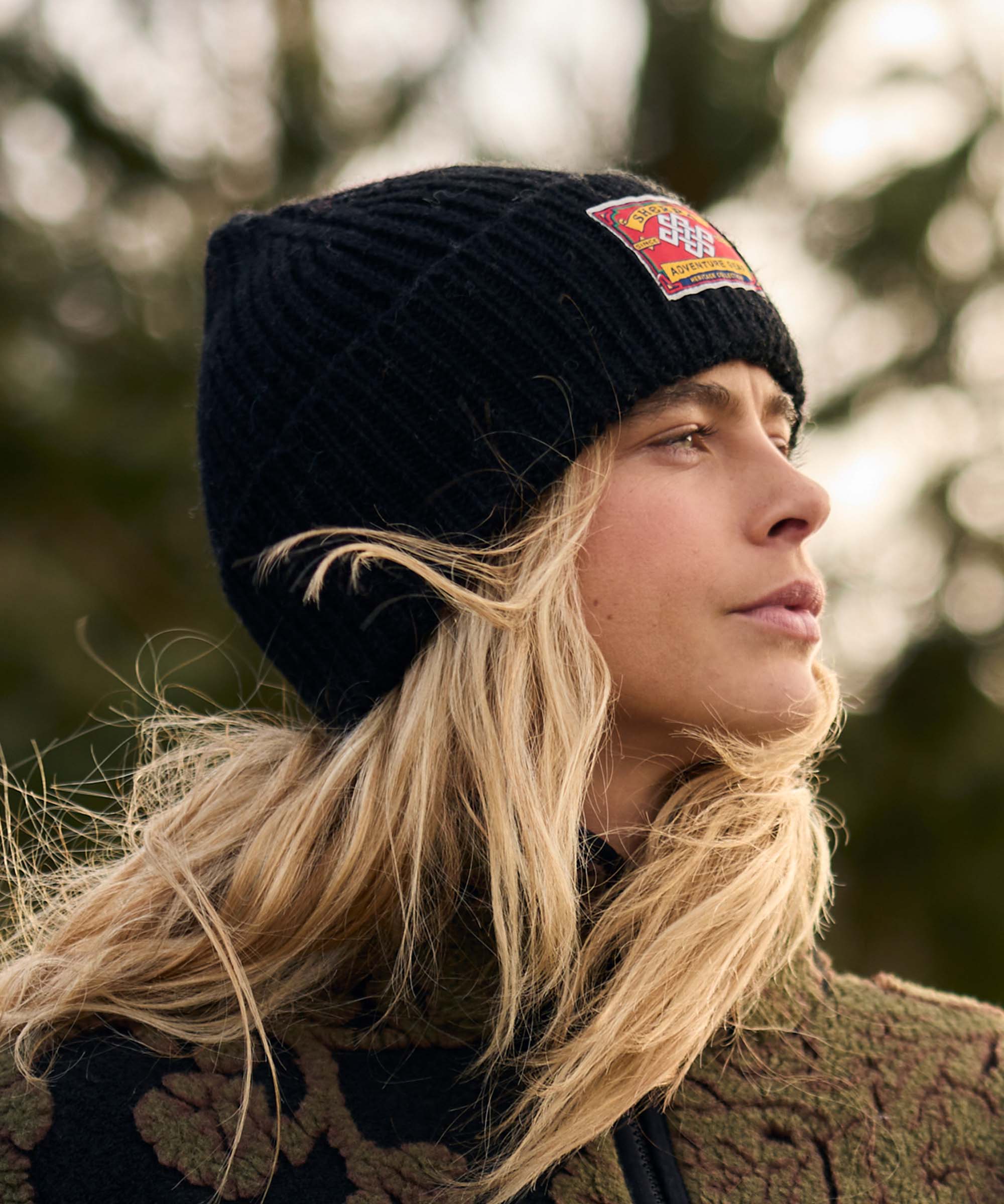 Woman standing among trees and greenery, looking toward the horizon as a breeze moves her hair. The outdoor setting feels quiet and reflective. She wears a black rib-knit beanie with a woven patch and a dark fleece jacket with an oversized floral pattern.